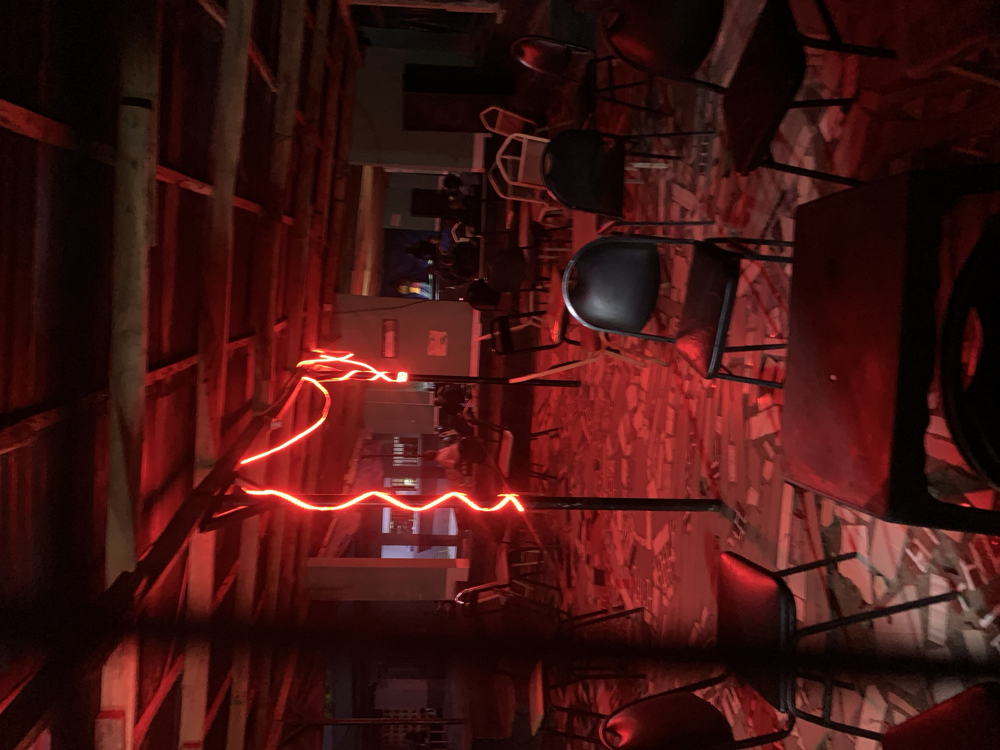 The image depicts the interior of a dimly lit establishment, possibly a bar or gathering place, in the city of New Stadium, Nigeria. The primary light source is a long, sinuous, red neon or LED strip that casts an intense red glow over the scene. The walls appear to be constructed of wood, perhaps exposed framing or paneling. Numerous metal chairs are scattered around the space, some arranged around tables. In the background, through an opening or doorway, are hints of other people and possibly windows in a building across the way. The floor appears to be made of tiles. The image has a slight tilt or is composed at an angle.