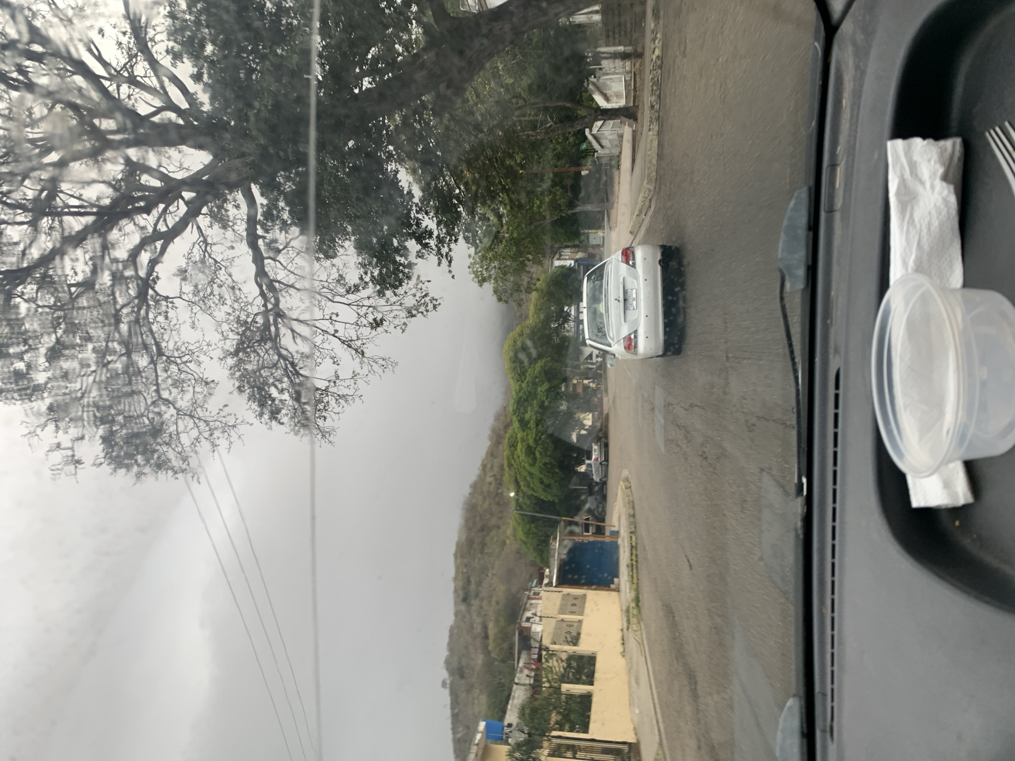 A view from inside a car, likely on the passenger side. The scene outside the window depicts a street in what is possibly La Asuncion, Venezuela, given the tropical foliage and architecture. A white car is driving ahead on a paved road. The environment is overcast and potentially rainy, as suggested by the water streaks on the glass. On the dashboard within the car, there is a clear plastic container, a white napkin, and a fork. Trees and buildings line the side of the road, and a hillside rises in the background. The image is slightly blurry and angled, giving it a casual, observational feel.