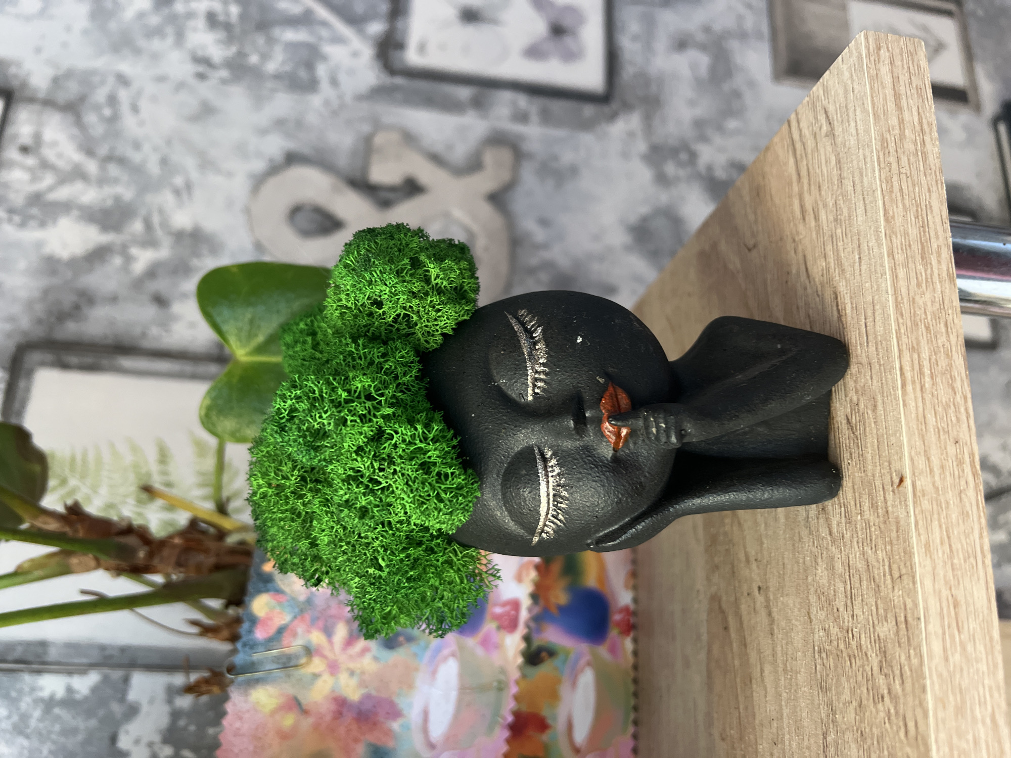 An indoor still life scene is captured, rotated 90 degrees clockwise, displaying a collection of decorative items. The central subject is a dark-colored, matte figurine, shaped like a human head with closed eyes, long silver eyelashes, and a small, slightly open mouth accented in orange or brown. A vibrant mass of textured, bright green material, resembling preserved moss, forms the "hair" on the figurine's head. A delicate silver fern-like pattern is visible on the forehead of the figurine in one of the crop hints.
To the left of the figurine, partially visible, is a green leafy plant with visible stems and roots, possibly in a small pot. Below the plant and extending towards the foreground, there's a piece of colorful fabric or paper adorned with a floral pattern in shades of pink, orange, blue, and green. A small gold-colored paperclip is attached to this material.
The figurine and plant are positioned on a light-colored wooden surface, likely a shelf or table, exhibiting a distinct wood grain pattern. The background consists of a light gray and white patterned wall, possibly wallpaper, featuring abstract shapes that resemble stylized letters or symbols (like a '2' and an '&' or 'S'), and a framed picture with a faint butterfly illustration.
There is no discernible activity taking place, as the image depicts a static arrangement of objects. The lighting is soft and even, suggesting an indoor setting during daytime. No explicit text is visible in the image.