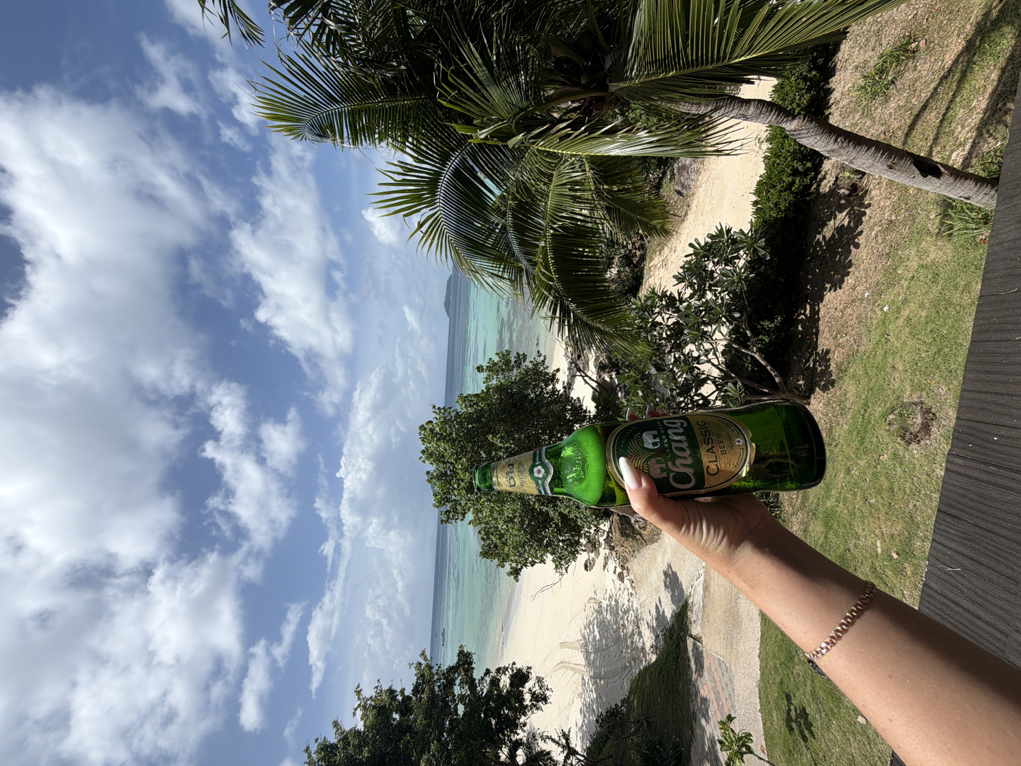 A hand with manicured fingernails holding a green bottle of Chang beer is in the foreground, positioned on the right side of the image. The hand wears a thin, rose-gold bracelet. The background features a tropical beach scene with white sand, clear turquoise water, and lush green trees, including a prominent palm tree on the right. In the distance, a small island is visible on the horizon. The sky is a bright blue with scattered white clouds. The scene appears to be a view from a balcony or elevated vantage point overlooking Ao Nang, Thailand.