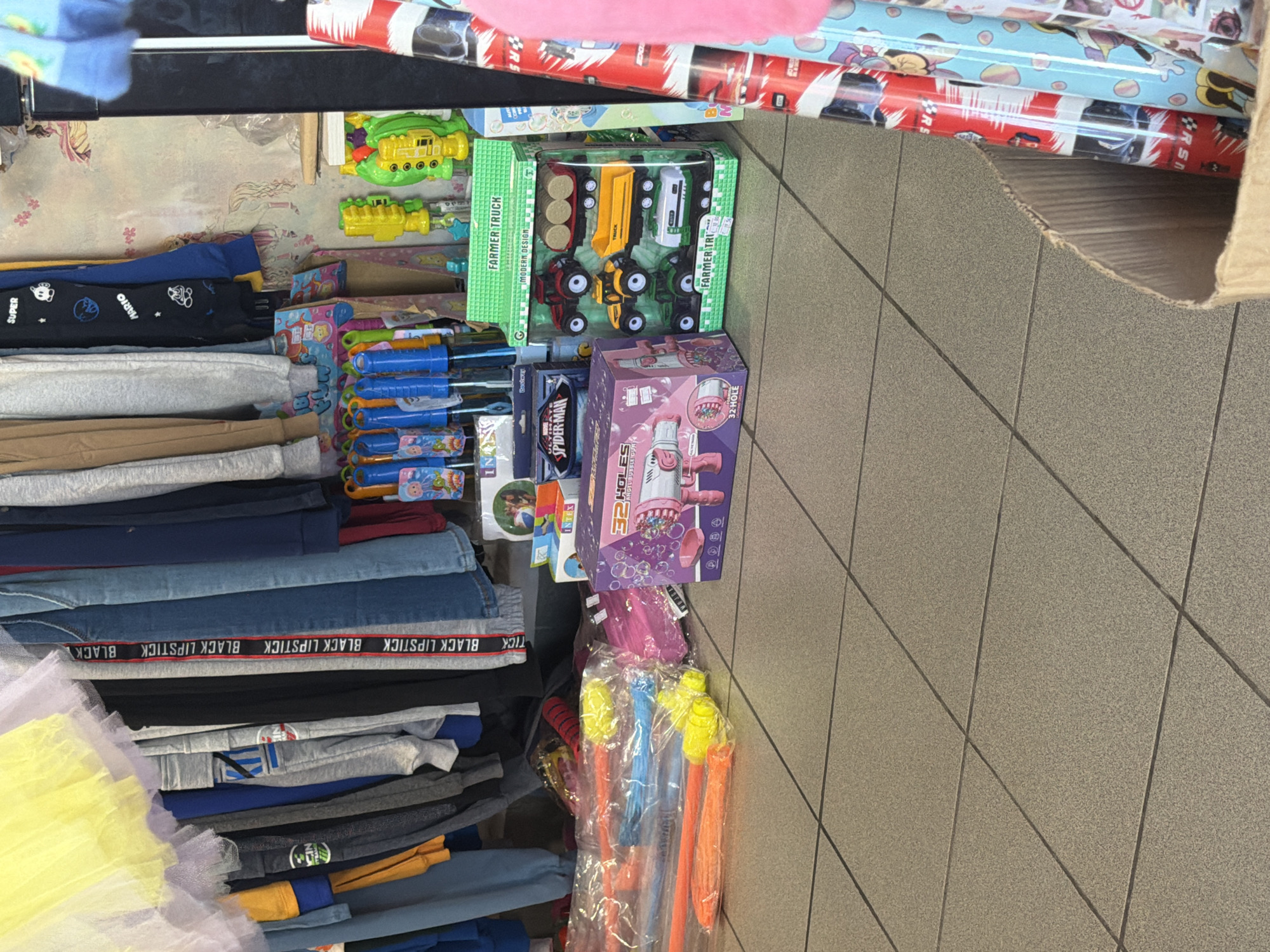 The image captures a store interior, likely a small retail establishment selling toys and clothing. The floor is tiled with light brown square tiles. To the left, a selection of children's clothing hangs on a rack, featuring various colors and patterns, including yellow tulle, blue denim, and grey sweatpants with visible branding like "BLACK LIPSTICK" and "SUPER MARIO".
In the foreground and mid-ground, several toy items are displayed. A box of what appears to be bubble guns sits to the right of the clothing rack. Further to the right, a green box containing a set of toy farm vehicles, including a tractor, a dump truck, and a tanker truck, is prominently displayed. Next to this, rolls of brightly colored wrapping paper are visible, some with cartoonish designs.
In the background, shelves are stocked with more toys and merchandise. The wall behind the merchandise has a patterned wallpaper featuring what appears to be unicorns and floral motifs. Two toy guns, one yellow and one green, are positioned on a shelf above the toy vehicles. A light blue object, possibly another toy, is also visible on the shelf. The overall impression is a well-stocked, somewhat cluttered retail environment aimed at children. There are no people visible in the image. The store is located in Ravno pole, Bulgaria.