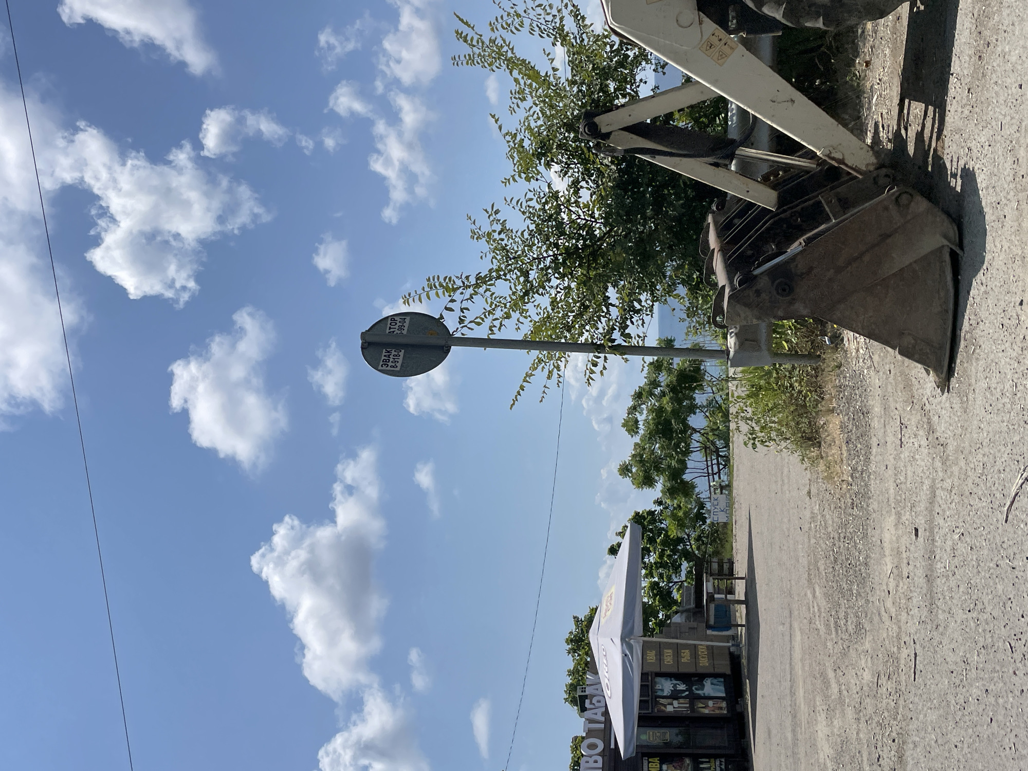 The image depicts an outdoor scene on a bright, sunny day with a blue sky dotted with fluffy white clouds. In the foreground, the bucket of a light-colored excavator is visible, partially obscuring a gravel road. To the left, a small kiosk or stand with a white awning is set up, displaying various items and signage, some of which appears to be in Russian. Behind the kiosk and to the right, green trees and foliage line the background, with a glimpse of the blue sea and horizon visible further in the distance. A metal pole with a circular sign attached is also present, along with a guardrail. The overall impression is of a roadside stop or small business in a coastal or seaside location. The city of Dzhubga, Russia is known for its Black Sea coast and is a popular tourist destination, fitting the visual elements of this image.