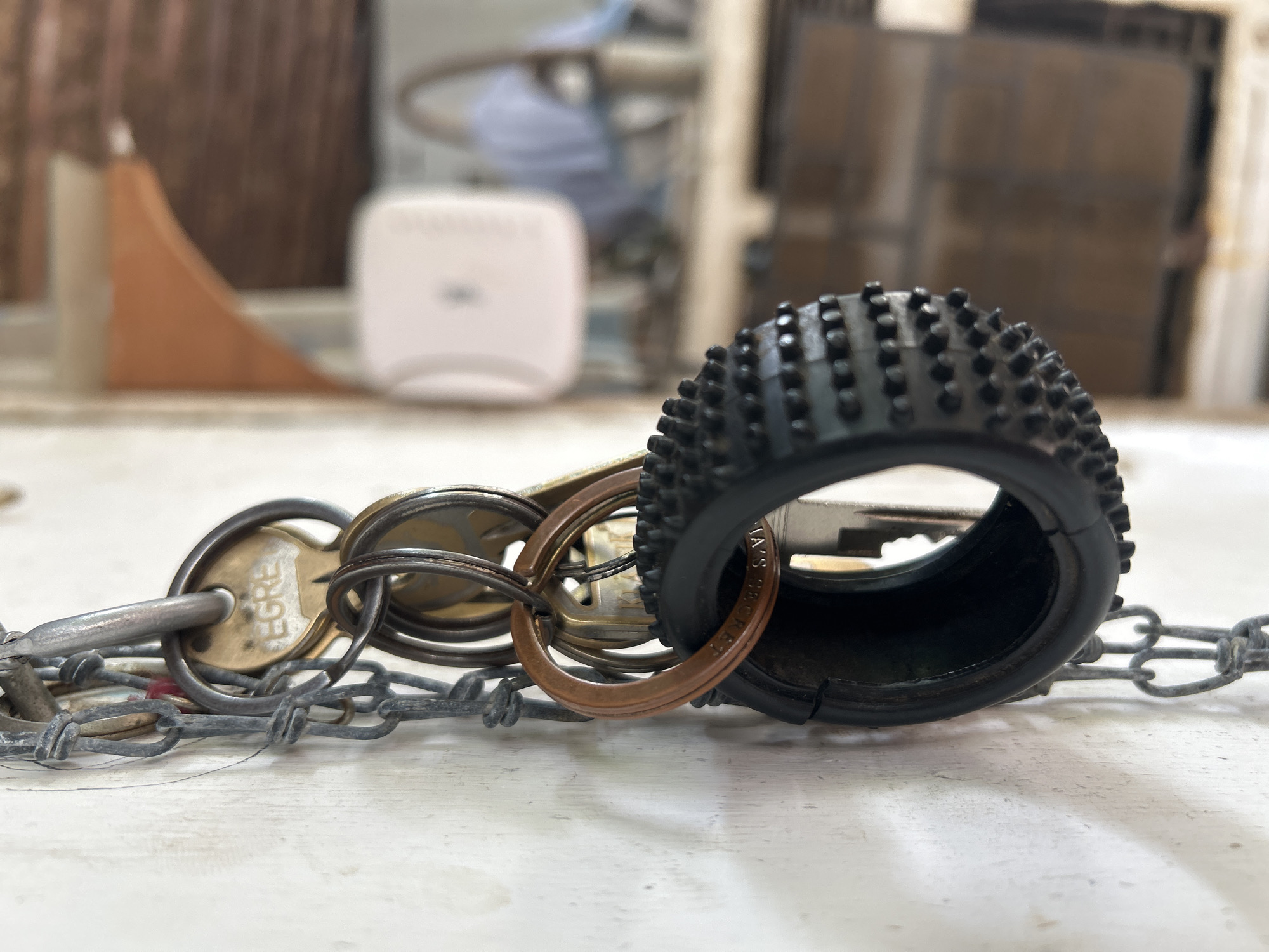 In Santa Ana, Venezuela, a close-up shot focuses on a cluster of keys hanging from a metal chain against a textured, off-white wall. The keys are attached to various keyrings, including one made of brass with the engraving "EGRE" visible and another of a copper-like metal. A prominent black, rubberized keychain resembling a tire with raised studs is also present. The background is blurred, showing hints of an indoor environment with what appears to be wooden shelving, possibly in a workshop or storage area. The lighting suggests an overcast or indoor setting.