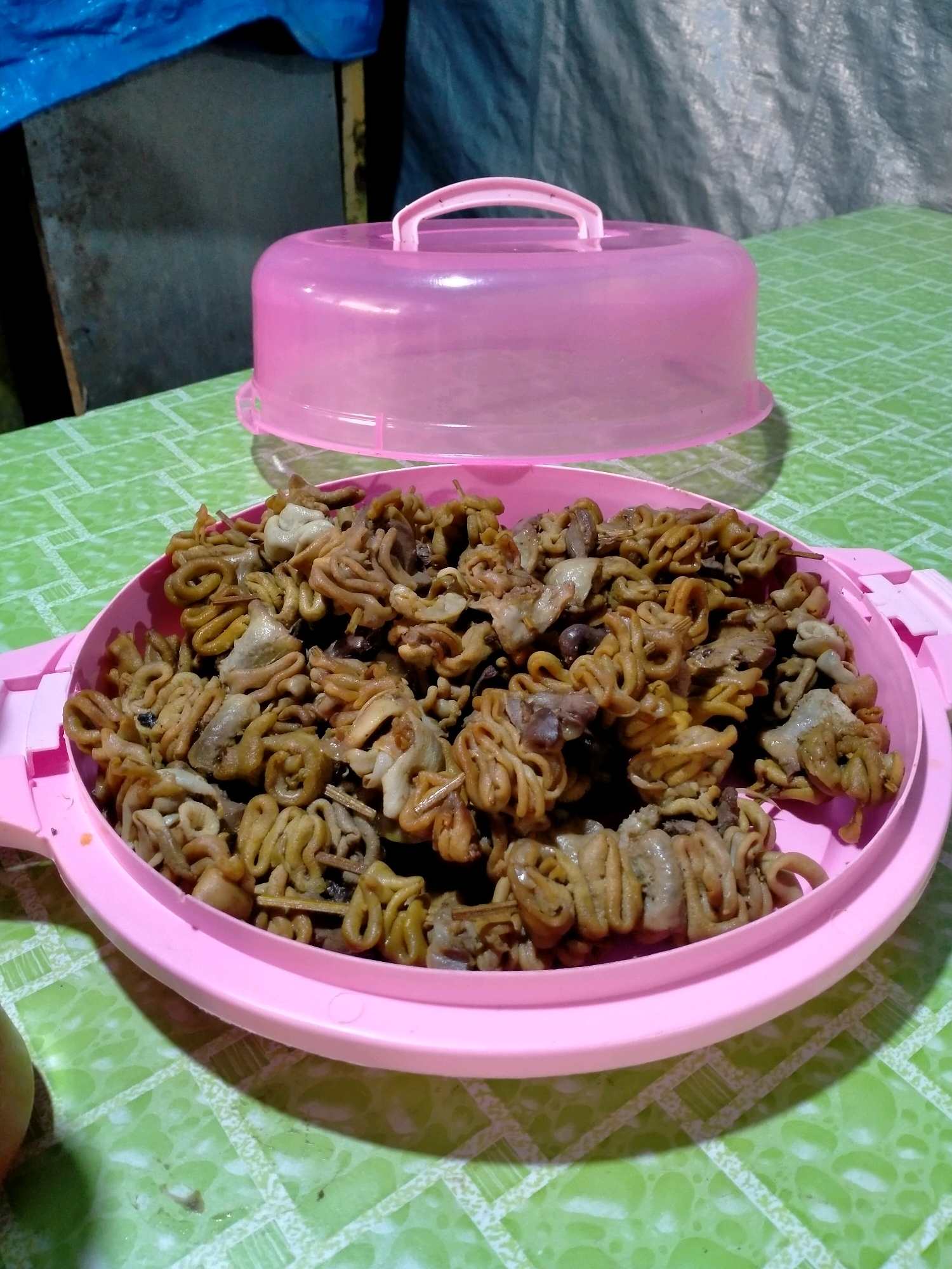A pink plastic serving dish filled with cooked intestines is on a green, patterned tablecloth. The dish has pink handles on either side and a pink plastic lid with a pink handle is propped open behind it. The intestines appear to be stewed or fried and are coiled and layered throughout the dish. The background shows a blurry blue tarp and a dark object. The scene is likely indoors, possibly in a home or a street food stall in Batu, Indonesia.