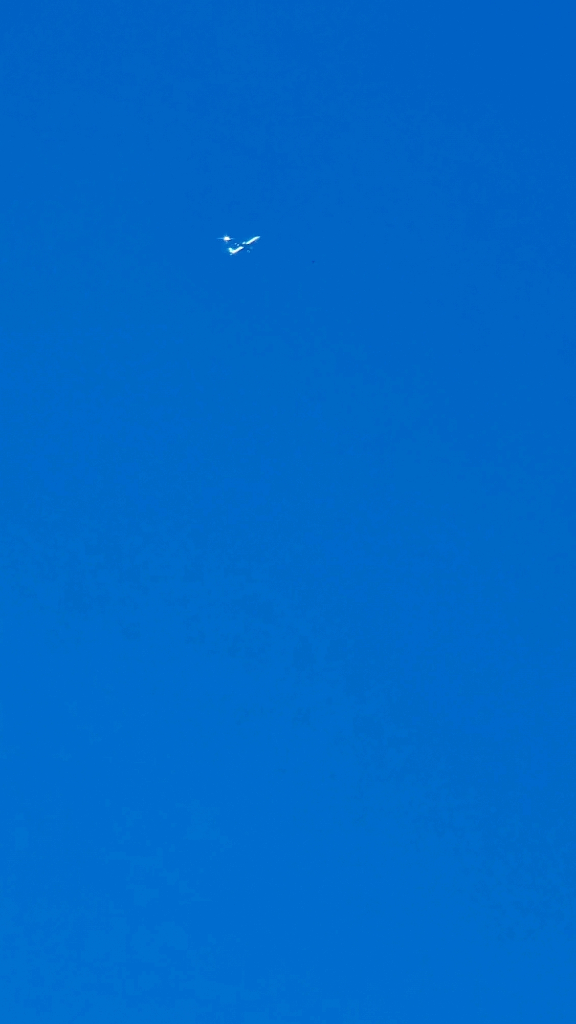 An airplane is visible against a clear blue sky. The airplane is small and white, with its wings extended. It appears to be a commercial jet. The image is taken from a low angle, looking up at the sky. There are no other objects or people visible in the image. The location of Llocalou, Spain, is not evident in the image.
