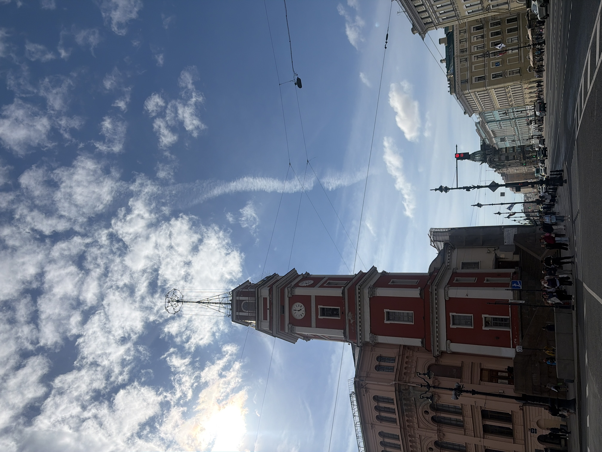 This image captures a view of the Peter and Paul Fortress's bell tower in Saint Petersburg, Russia, under a bright blue sky with scattered clouds and the sun shining through. The red and white bell tower stands tall, featuring clock faces and a distinctive spire topped with a globe and a star. Adjacent to the tower is a pale pink building with arched windows. Below, a street scene unfolds with people walking on sidewalks, crossing roads, and a few cars on the street. Overhead, power lines and a lamppost are visible. The perspective is from street level, looking up towards the tower and the sky, with more urban architecture lining the street in the distance.
