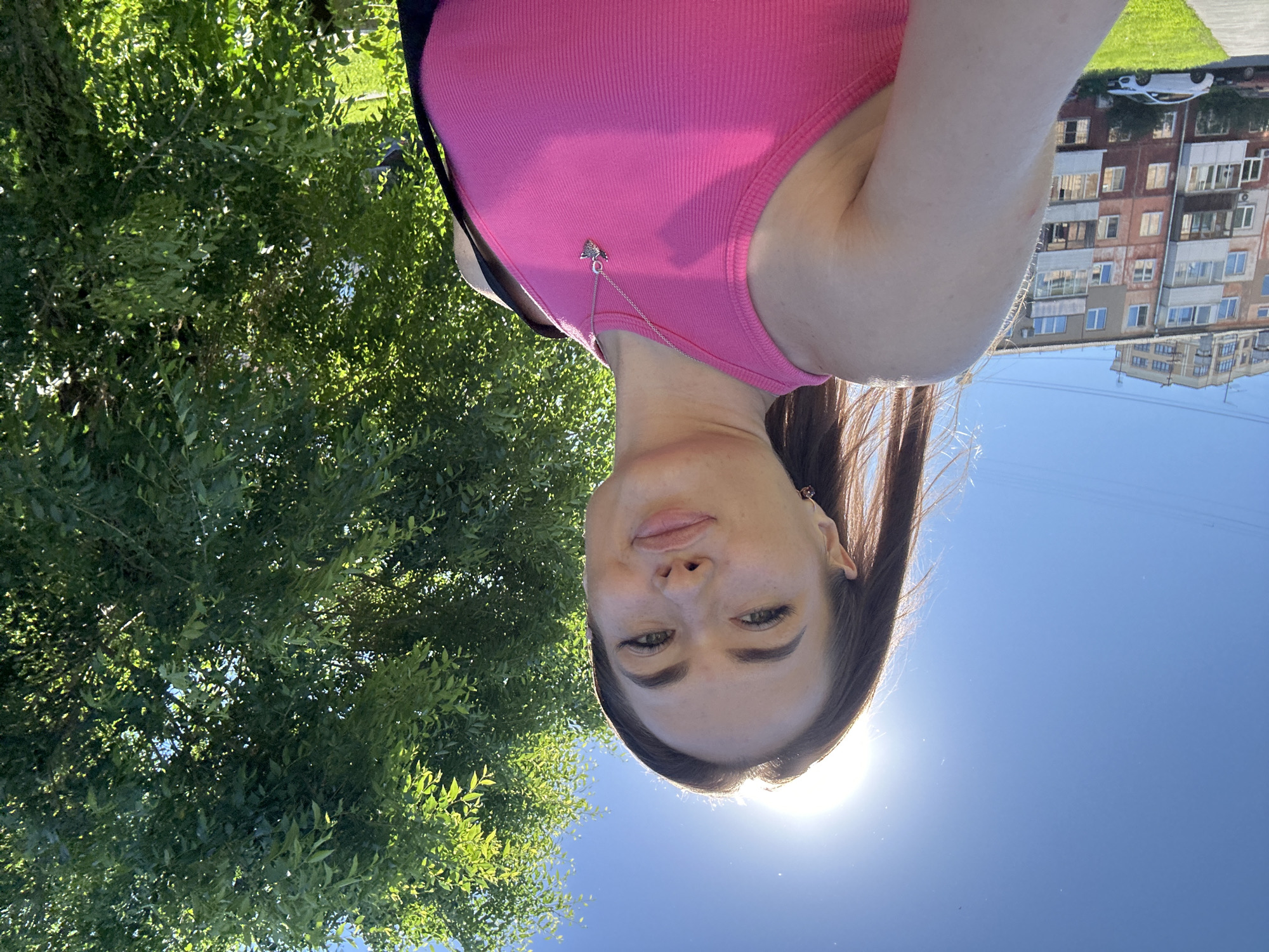 In an outdoor, low-angle, full-body selfie, a young woman stands in front of a leafy green tree and a residential building. The sun is high in the sky and flares over her left shoulder. She has long, dark brown hair and is wearing a bright pink ribbed tank top and a silver pendant necklace. Her eyes are green, and her lips are slightly pursed. To her left, a multi-story apartment building with numerous windows and balconies stands under a clear blue sky. A white car is parked on the street in front of the building, and a patch of green grass is visible. The scene is brightly lit by natural sunlight.