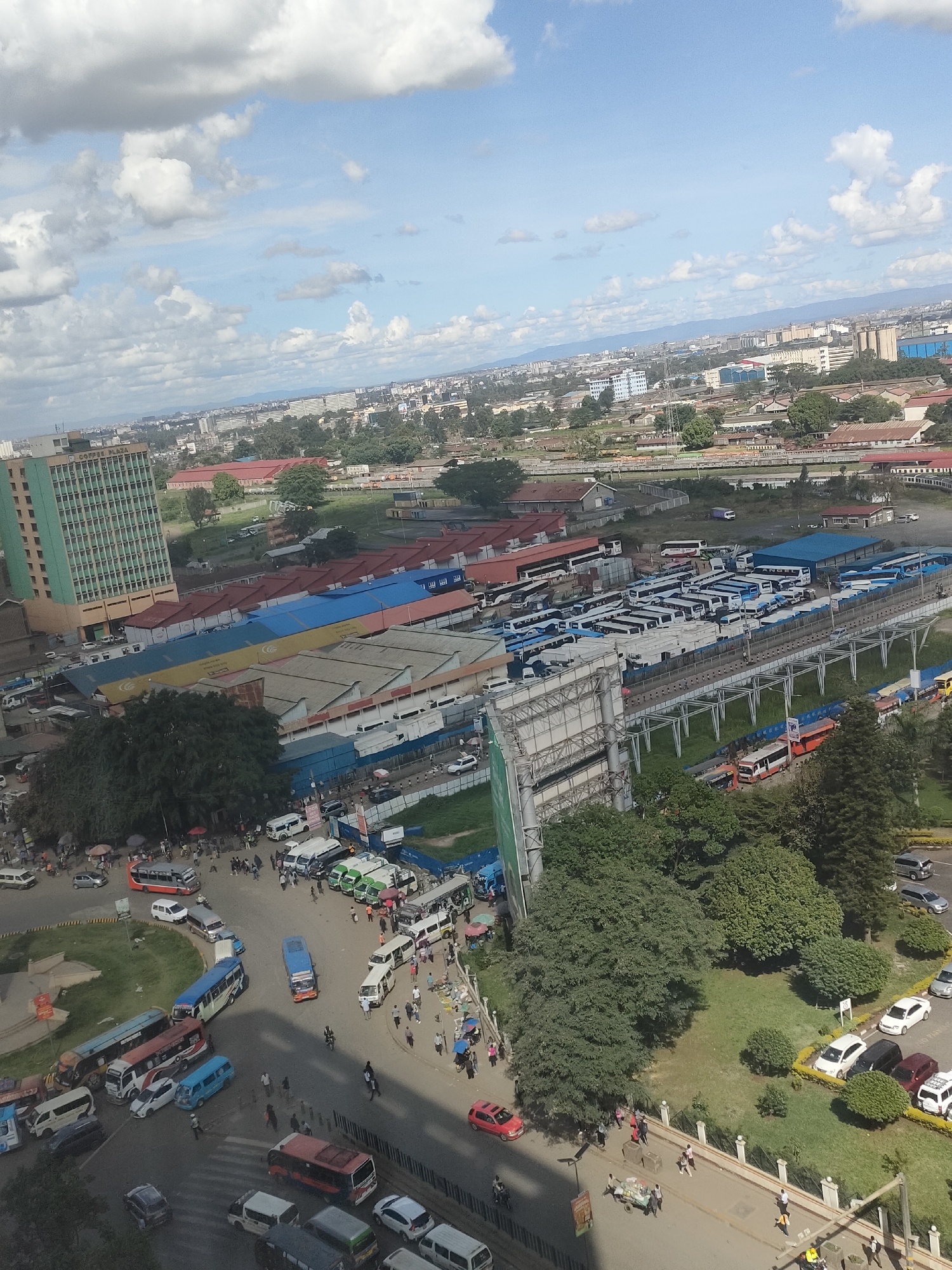 This is an elevated view of a bustling area in Nairobi, Kenya. In the foreground, a roundabout teems with traffic consisting of buses, matatus (minibuses), cars, and motorcycles. Pedestrians are visible crossing roads and navigating the roundabout. Beyond the roundabout, there are lines of parked buses under a metal roof structure with a railway line adjacent to it. There are large industrial buildings with blue and red roofs. In the mid-ground, green spaces with trees, including a parking lot, give way to urban sprawl with buildings stretching into the distance. The sky is mostly blue with scattered white clouds.