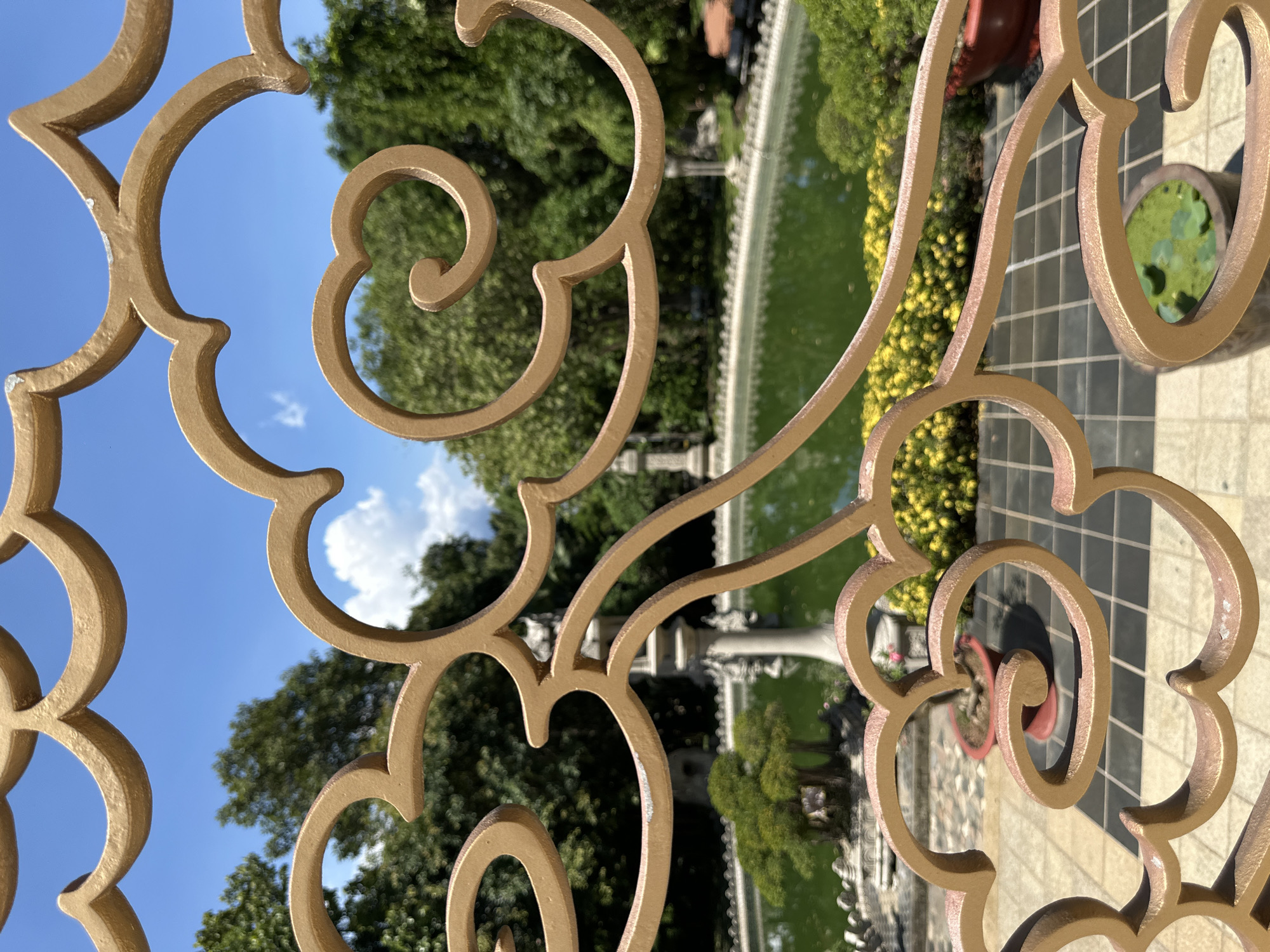 A close-up shot of an ornate, gold-colored metal fence partially obscures a view of a park in Dĩ An, Vietnam. The fence's design incorporates curls and floral motifs. Through the fence, the background reveals a rectangular pond or water feature, a stone fountain, manicured bushes, trees, a stone-tiled pathway or patio. In the foreground, a potted plant with green leaves is partially visible. The sky is blue with white clouds visible through the fence.