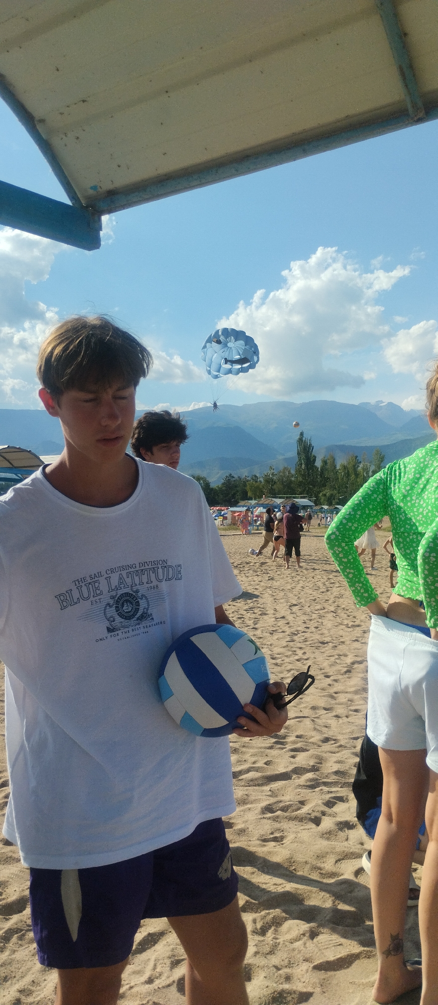 In Cholpon-Ata, Kyrgyzstan, on a sandy beach, a young man in a white t-shirt and purple shorts stands holding a blue and white volleyball and sunglasses. The t-shirt has a graphic that reads "THE SAIL CRUISING DIVISION BLUE LATITUDE EST 1988 ONLY FOR THE BEST SEAFARERS". In the background, a paraglider is suspended in the sky, with mountains and trees visible behind it. Other people are scattered on the beach, some appearing to be engaged in activities. The sky is blue with scattered clouds, and the scene is illuminated by sunlight.