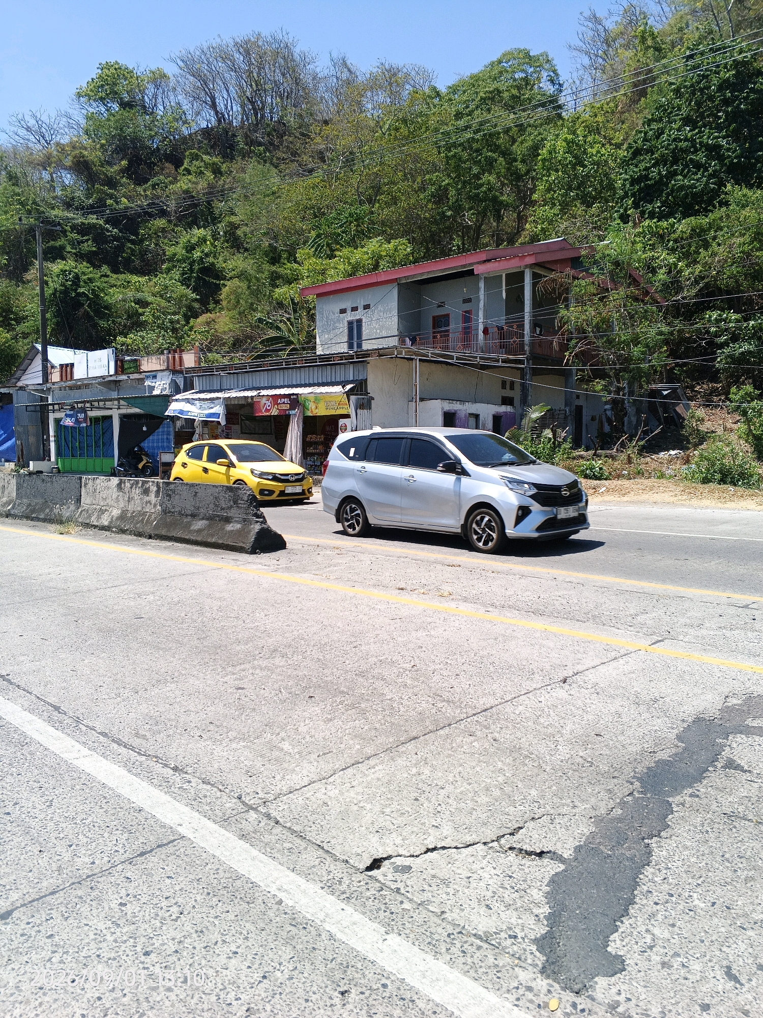 This image captures a street scene in Kupa, Indonesia, on a bright, sunny day. A silver hatchback is traveling on a paved road, with a yellow hatchback in the adjacent lane. The road is marked with white and yellow lines. A concrete barrier separates the road from a row of small shops and buildings situated on a sloping hillside covered in dense green foliage. Power lines are visible across the scene, strung between utility poles. The buildings appear to be a mix of commercial establishments and residential structures, with some having red-tiled roofs. The overall environment suggests a moderately developed area with a natural backdrop.