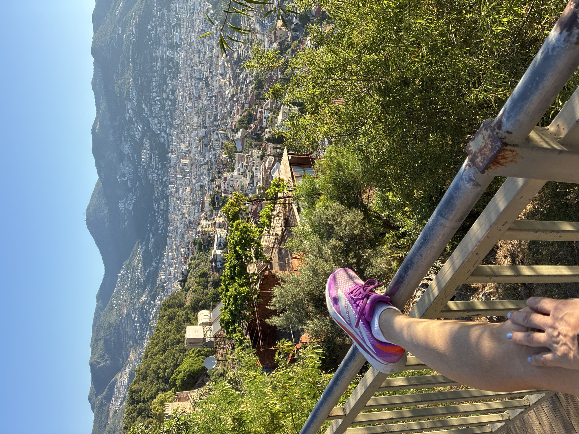 A first-person perspective shot shows a person's leg resting on a metal railing, with a sprawling cityscape of Alanya, Turkey, in the background. The leg is clad in a purple and orange athletic shoe, white socks, and skin with visible texture. The hand is resting on the thigh, with light blue nail polish on the fingernails.
The cityscape extends up a hillside, with numerous buildings and roads visible. Lush green trees and vegetation are scattered throughout the scene, with a clear blue sky above. The railing appears weathered, with some rust visible. The image captures a moment of relaxation or contemplation with a panoramic view of the city and its mountainous surroundings.