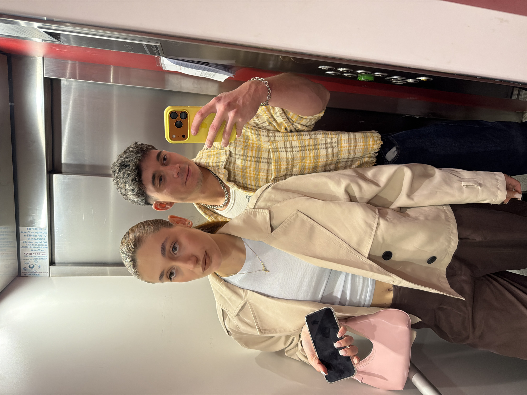 Two people, a man and a woman, are taking a selfie in what appears to be an elevator. The man, wearing a yellow plaid shirt and a chain necklace, is holding a yellow phone and looking at the camera. The woman, dressed in a beige trench coat, white top, and brown pants, is holding a black phone and a pink purse. The elevator has metal walls with a panel of buttons above them. A sign with text is visible on the wall. The location is likely Sofia, Bulgaria, based on the Cyrillic text on the sign.