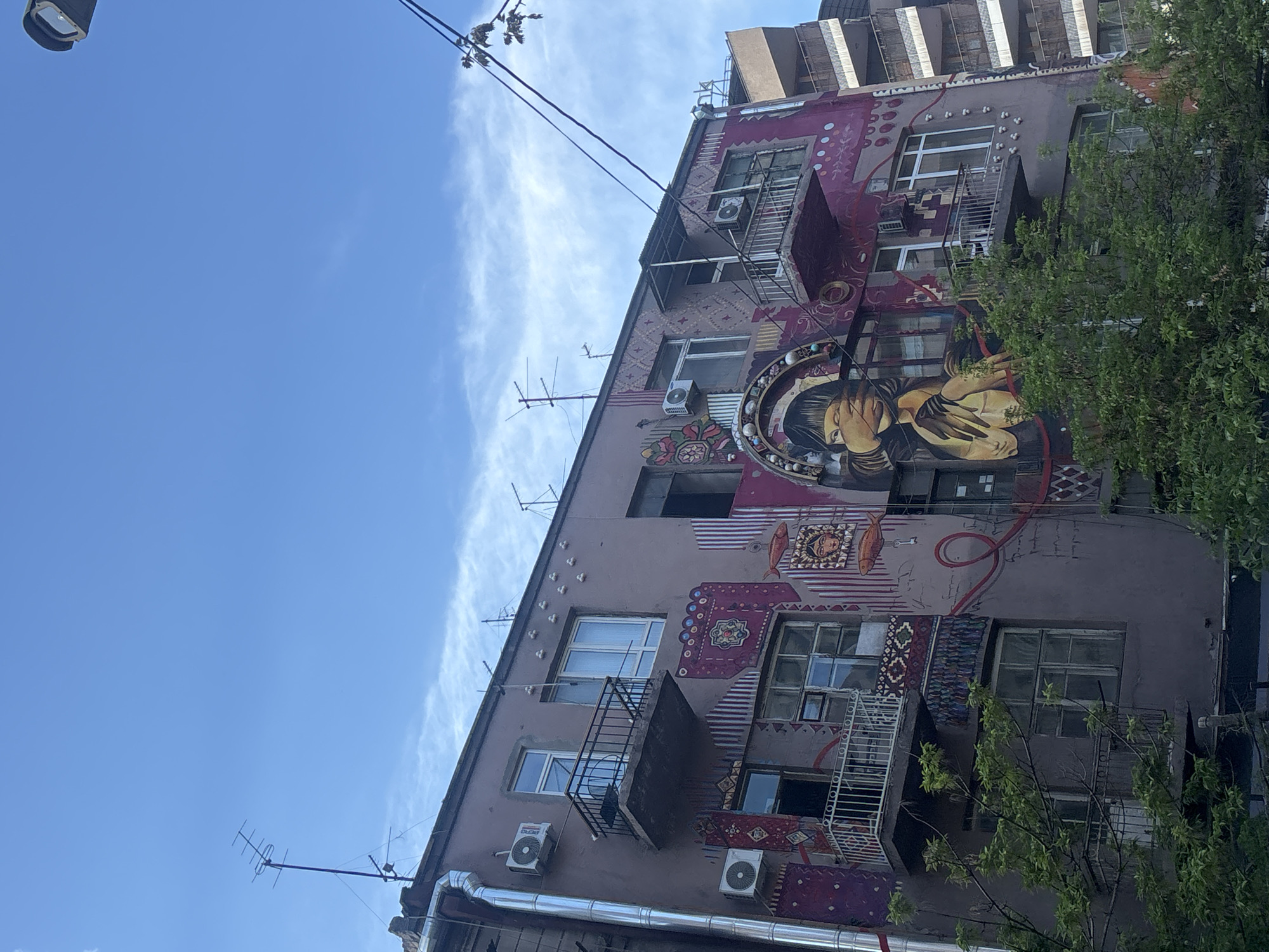 The image shows a multi-story building in Yerevan, Armenia. The building's facade is decorated with a large mural of a woman with her arms crossed in front of her chest. The mural is surrounded by geometric designs. There are balconies and windows on the building. Air conditioning units are attached to the exterior walls. The sky is blue with wispy clouds. Tree branches partially obscure the building on the right side. Power lines are visible in the sky.