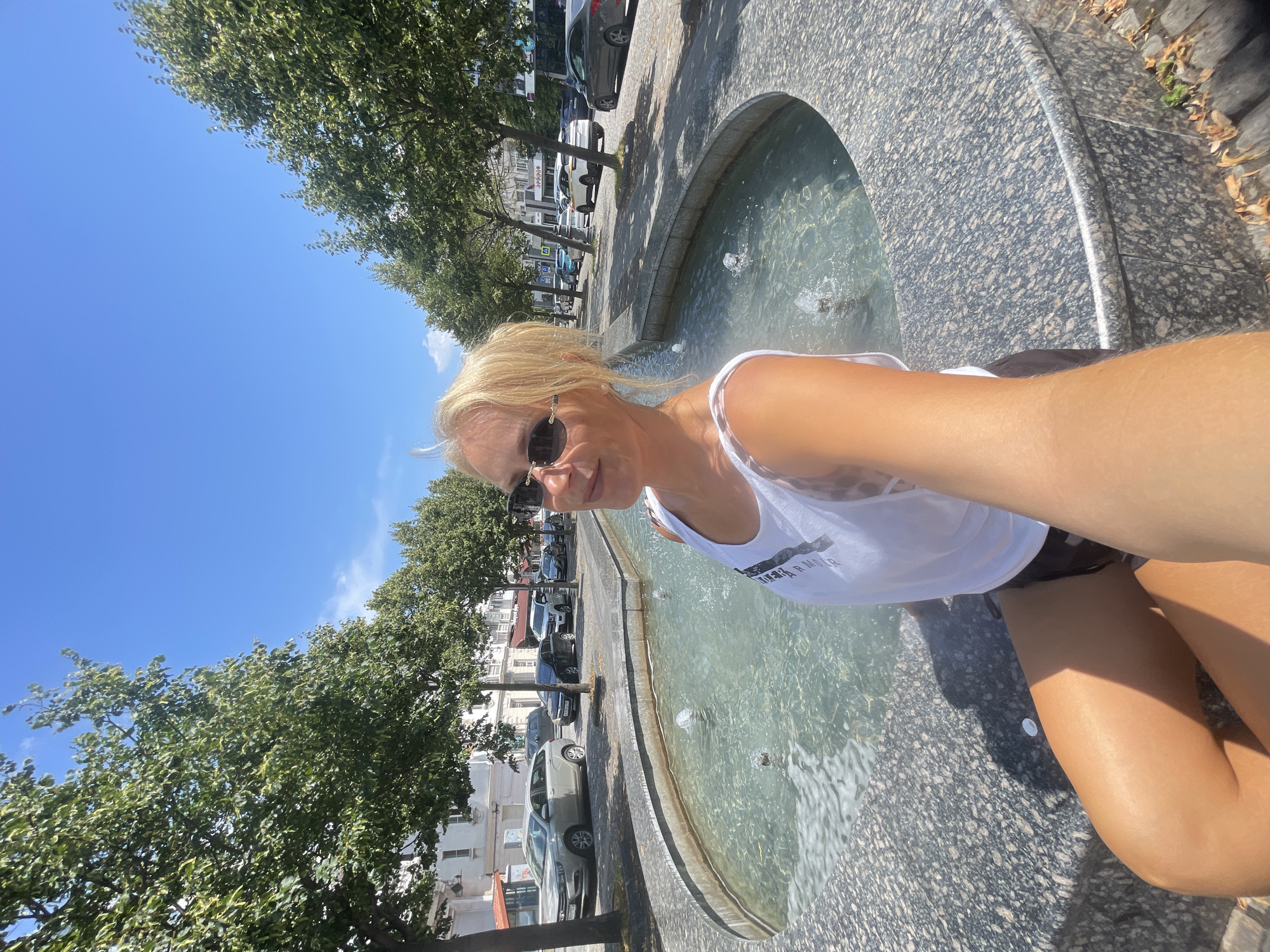 An outdoor selfie shows a blonde woman in sunglasses and a white tank top sitting on the edge of a decorative fountain. The fountain has a granite surround and water jets. Behind her, a tree-lined street is visible with parked cars and buildings. The sky is clear and blue. The scene appears to be in the city of Novorossiysk, Russia.