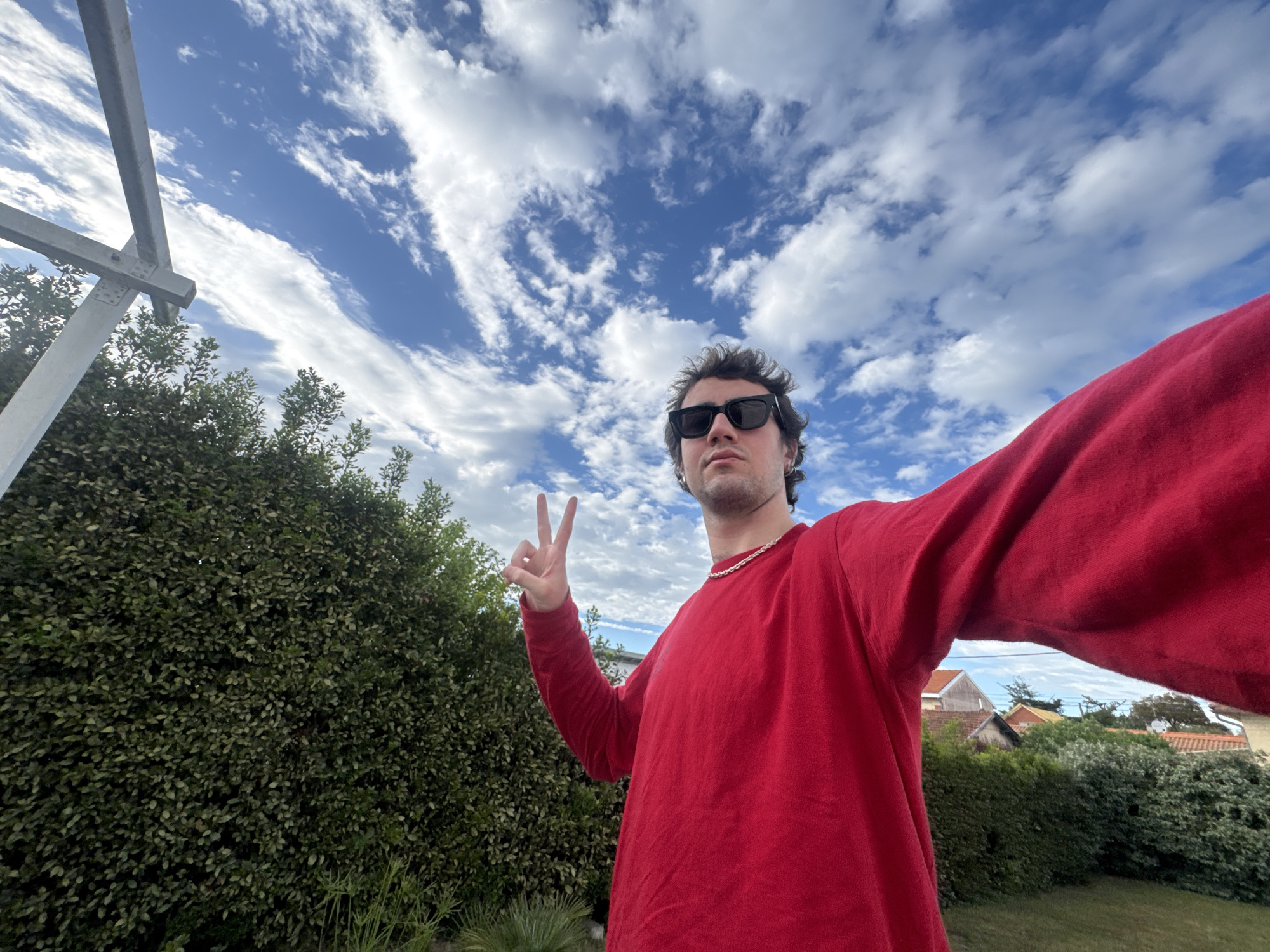 A selfie captures a young man in an outdoor setting. He wears a red long-sleeved shirt, a silver chain, and dark sunglasses. His curly hair is brown. He makes a peace sign with his left hand. The background reveals a vibrant blue sky filled with white clouds. Behind him are green bushes and a glimpse of a residential area, including houses and more greenery. A gray metal structure is partially visible on the left side of the frame. Based on the residential architecture, greenery, and visible sky conditions, the location can be identified as Soulac-sur-Mer, France.