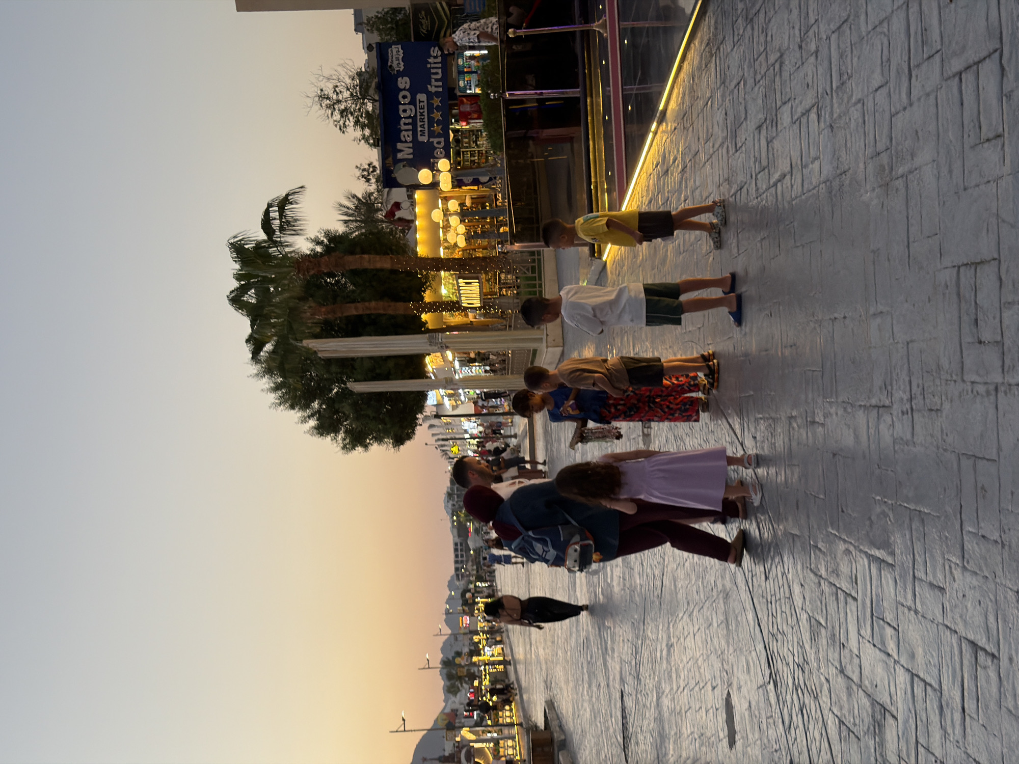 The image depicts a pedestrian promenade in Sharm El Sheikh, Egypt, during twilight. A group of people, including adults and children, are gathered on a paved walkway lined with shops and palm trees. The sky is a soft gradient of orange and pale blue, indicating the end of the day. In the background, lights from storefronts illuminate the scene, and mountains are visible in the distance. A prominent sign for "Mangos MARKET" with "fresh fruits" is visible on a building. The individuals are casually dressed, suggesting a relaxed, holiday atmosphere. One adult, wearing a headscarf, is interacting with several children. Another woman in a black dress is walking away from the camera further down the promenade. The paving stones are laid in a herringbone pattern.