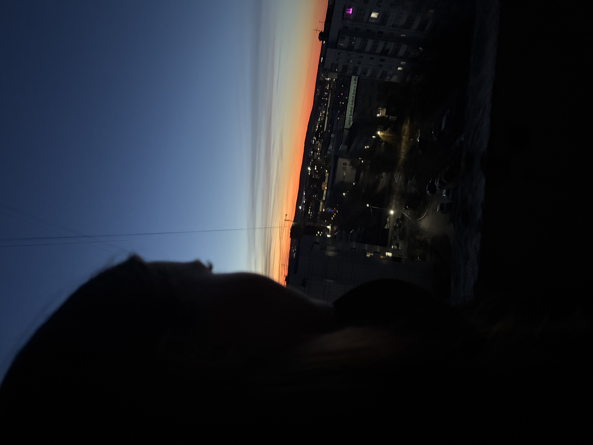 The image shows a cityscape at dusk or dawn in what is likely the city of Chaikovsky, Russia. A silhouette of a person, partially visible, is in the foreground. Behind the person, a gradient sky transitions from dark blue to orange, indicating the horizon. Below the horizon, a mix of residential and commercial buildings are visible, with lights illuminating windows and streets. The scene includes streets, trees, and potentially cars or other objects on the ground. The image is taken from a slightly elevated perspective, possibly a balcony or window.