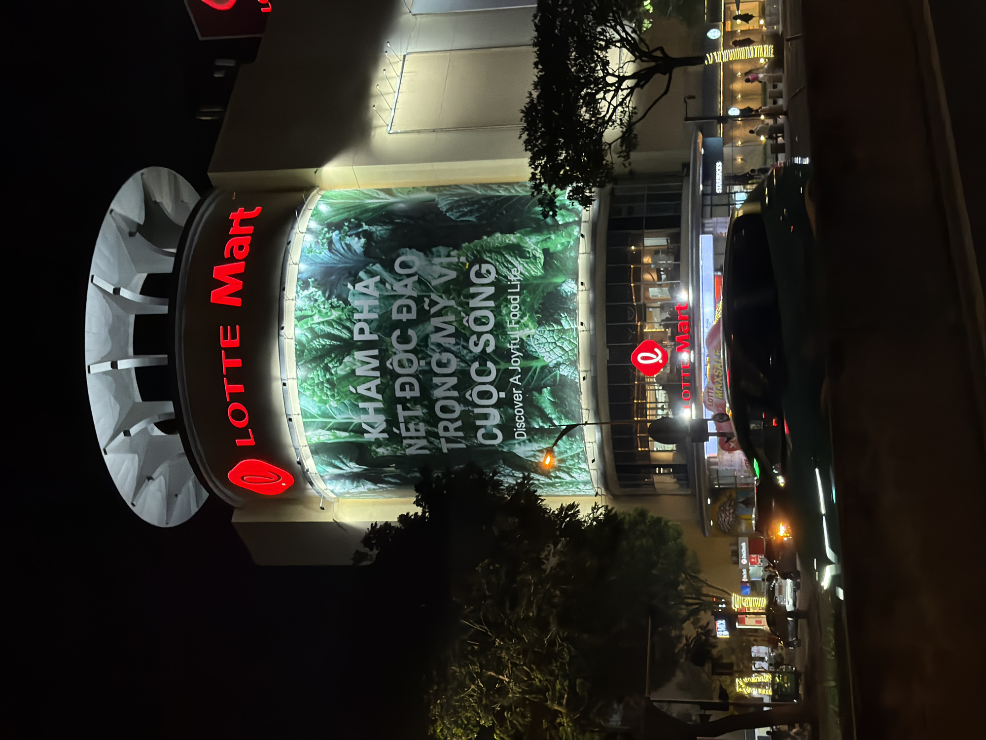 The image shows a Lotte Mart department store in Da Nang, Vietnam at night. The store has a large, illuminated sign that reads "LOTTE Mart" in red letters. A large banner on the side of the building displays an image of fresh produce with Vietnamese text that translates to "Discover the unique taste of a joyful food life". The street in front of the store is busy with traffic, including cars and motorcycles. Pedestrians can be seen walking on the sidewalk. The scene is illuminated by the store's lights, streetlights, and car headlights, creating a vibrant night-time atmosphere.