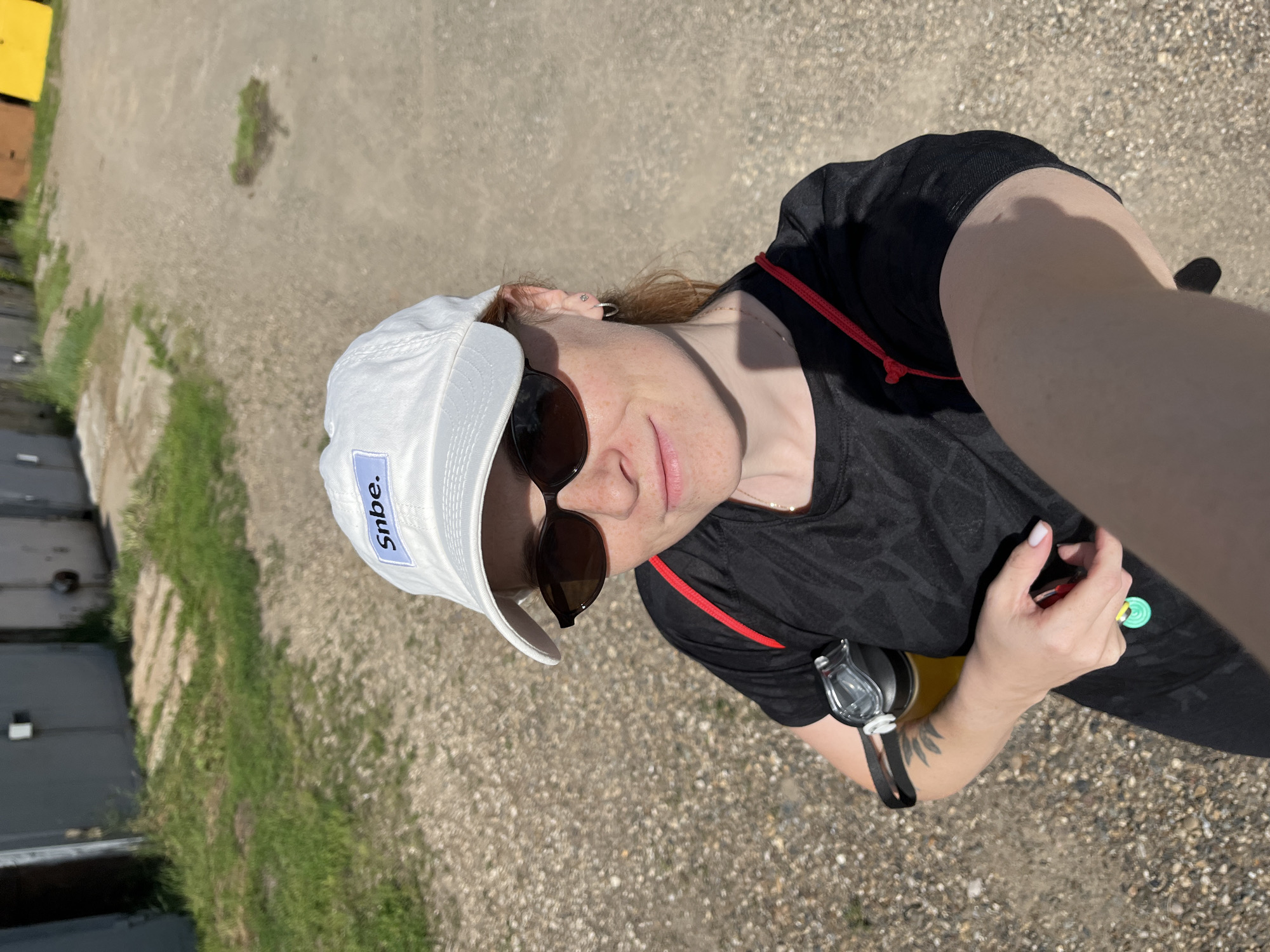 An outdoor, eye-level, close-up, selfie of a young, fair-skinned woman wearing a white baseball cap with a blue patch that says "Snbe.", dark-rimmed sunglasses, and a black t-shirt with a red strap over her shoulder. The woman has red hair and freckles. She is holding a water bottle with a yellow liquid in her left hand and a set of keys in her right hand. The background consists of a gravel path, some green grass, and a row of gray garage doors. The scene appears to be in a residential area of Chaikovsky, Russia, possibly near a garage cooperative. The lighting suggests it is daytime, likely sunny.