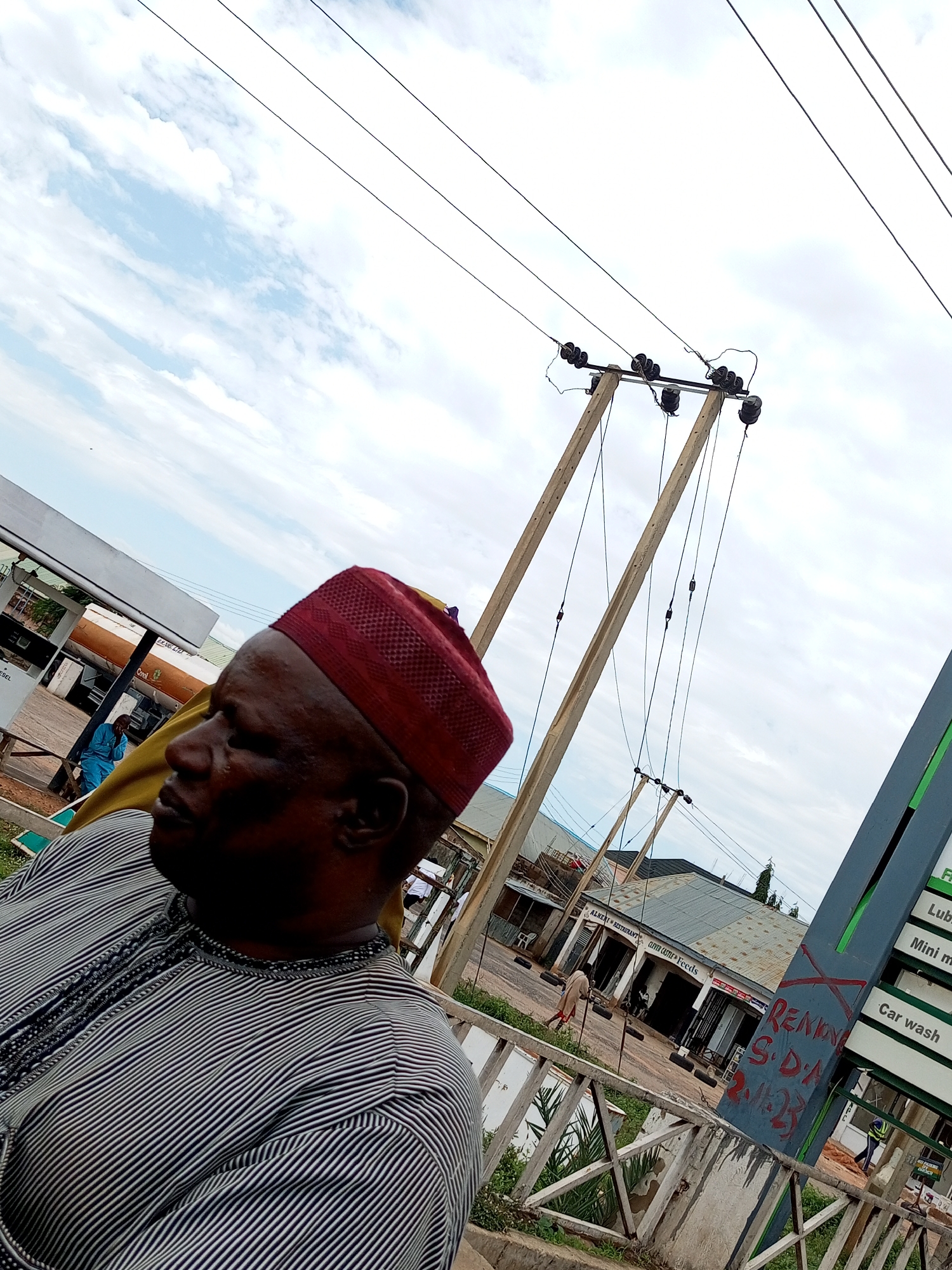 This is a portrait photograph taken outdoors in Gombe, Nigeria. The subject, a middle-aged man wearing a red cap and a striped traditional garment, is positioned in the foreground, facing left and looking downwards. In the background, a gas station is visible with a man in a blue shirt standing near a fuel pump. The sky above is filled with clouds. The image also shows overhead power lines and a sign with the word "Car wash". The overall setting suggests a public space within the city.