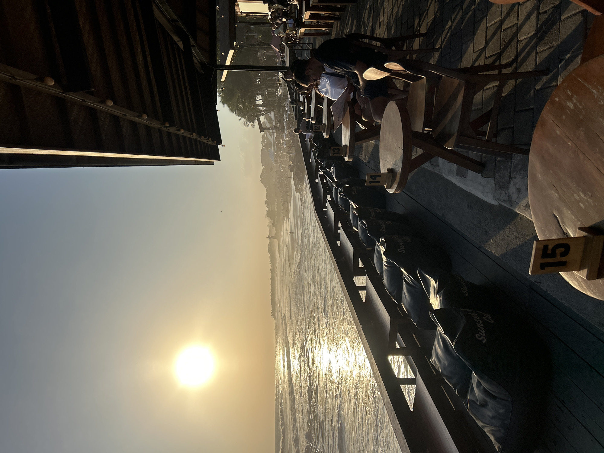 The image depicts an outdoor seating area of a cafe, likely "Sunset Cafe" as indicated by lettering on beanbag chairs, situated along a beach in North Kuta, Indonesia. The setting sun casts a warm golden glow over the scene, with the sun itself positioned prominently in the upper left quadrant. The ocean stretches out to the horizon, featuring moderate waves and a reflective, sunlit surface.
In the foreground, there are rows of dark beanbag chairs facing the ocean, with wooden benches behind them. Several wooden tables and chairs are also present, with some patrons seated at them. One couple is visible at a table, with the man looking at a menu. The flooring consists of wooden planks and paved stones.
Further back, the beach is populated with many people, umbrellas, and tents, suggesting a lively atmosphere. In the distance, palm trees and some structures are visible against the hazy sky. The cafe itself has a covered roof supported by wooden beams, with string lights hanging from it. The overall impression is one of a relaxed, scenic dining or lounging experience during sunset.
