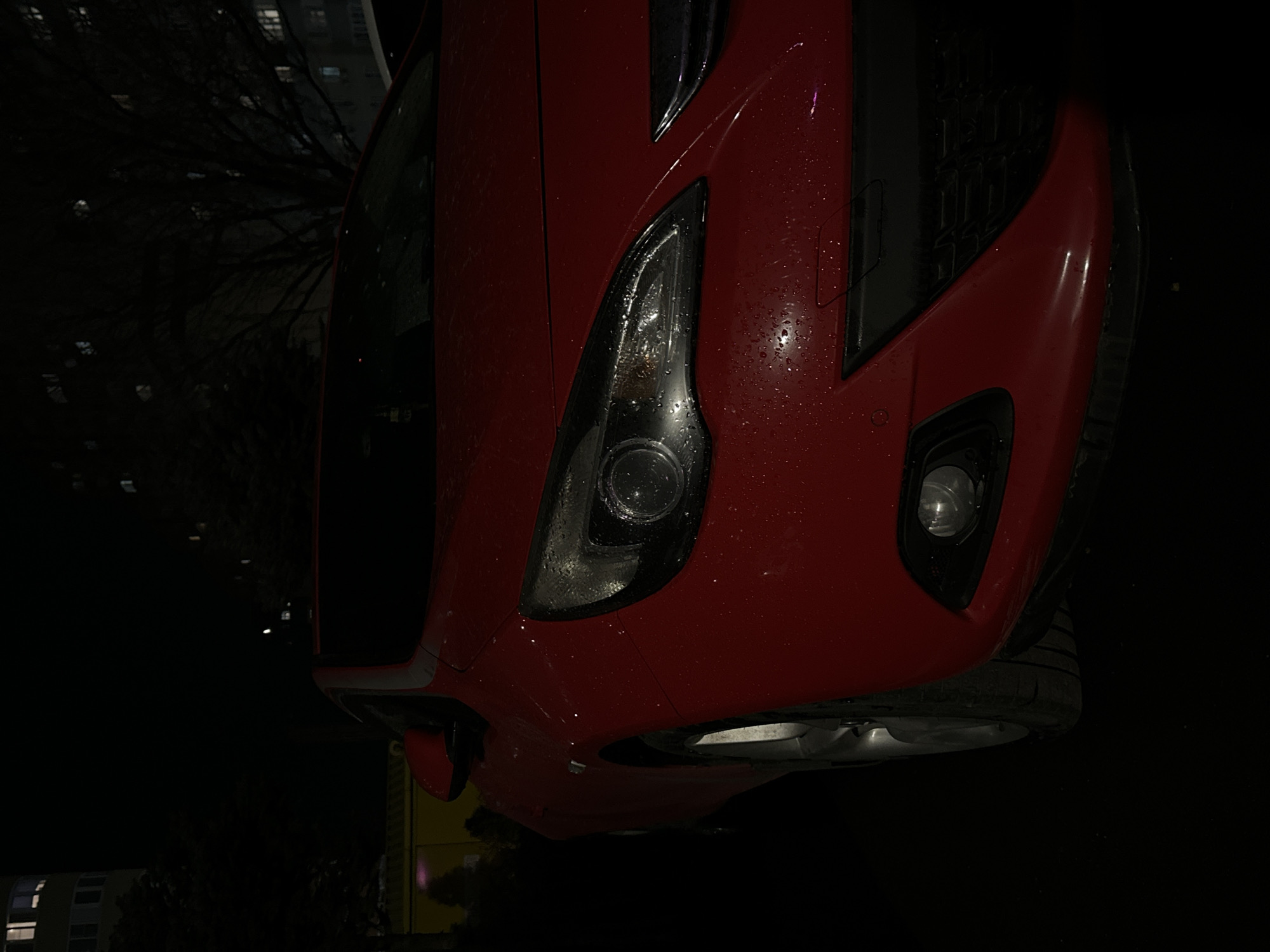 A close-up, low-angle shot depicts the front of a red car, possibly a hatchback, parked on a dark street. The vehicle is wet, with visible water droplets on the headlight and body. The headlight is on, and a small fog light is visible on the front bumper. A tire is partially visible near the lower right of the frame. The surrounding environment is dimly lit, and hints of buildings and trees are visible in the background. It's nighttime. The location, Ovoshchnoy, Russia, is unconfirmed from the visual data alone.