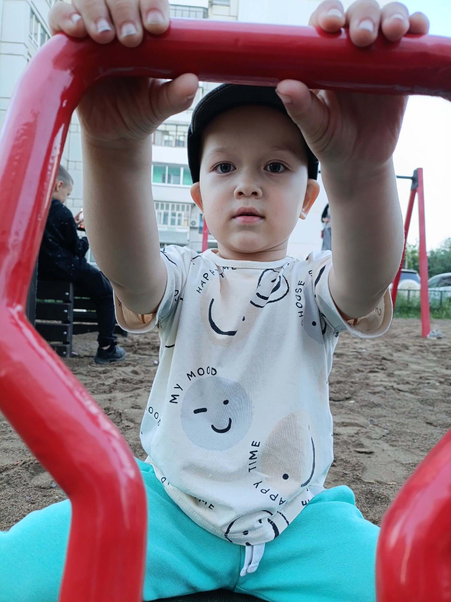 A close-up, eye-level medium shot captures a young boy wearing a black cap and a white t-shirt adorned with cartoon faces, sitting on a playground. His hands grip a bright red metal bar in the foreground. The boy's gaze is directed towards the camera, with a neutral expression. In the blurred background, another child, possibly older, is seated on a dark bench, looking at a mobile phone. The ground is covered with sand, typical of a playground, and a building with light-colored facades and windows is visible behind the children. The scene is likely situated in a residential area of Chaikovsky, Russia, given the architectural style of the building.