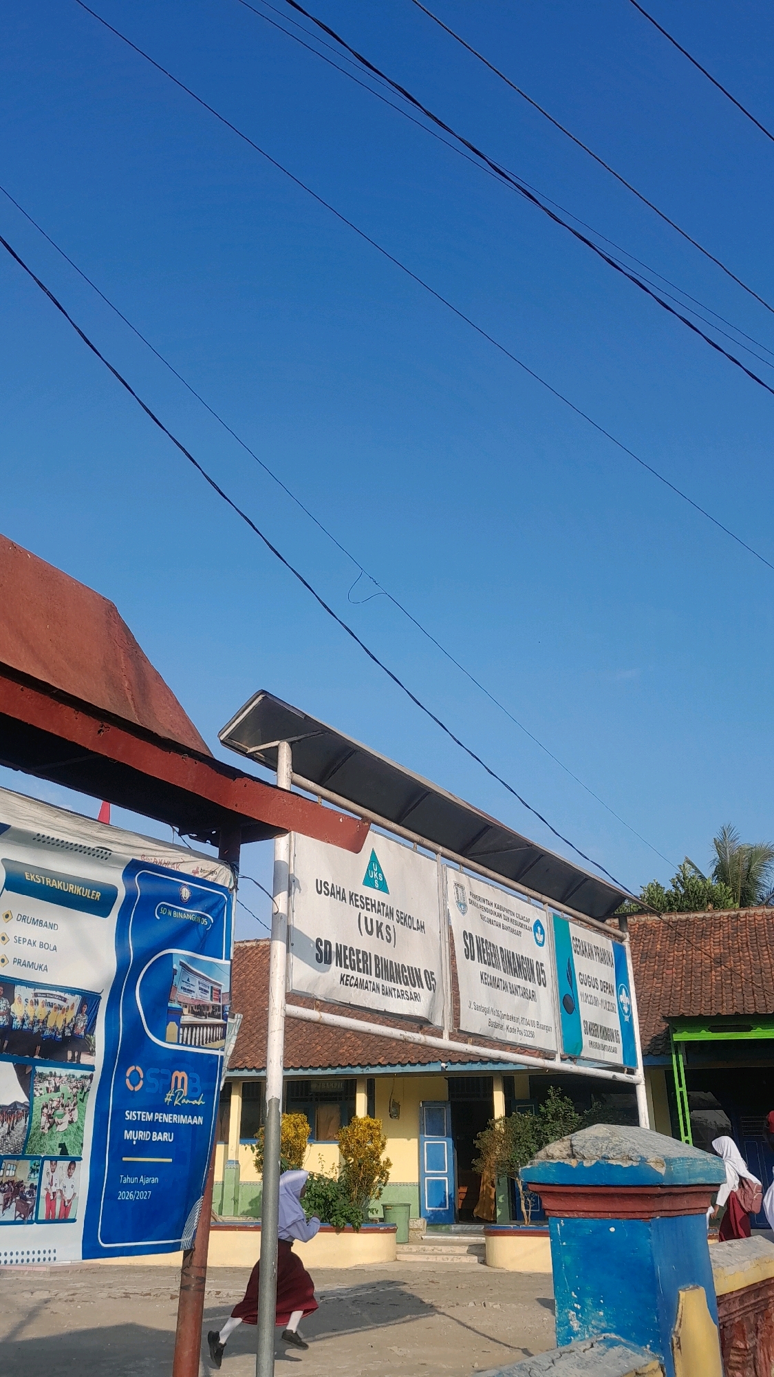 The image depicts a street scene in front of a school building. A blue sky with several power lines spans the upper portion of the frame. In the foreground, a child in a school uniform, a white hijab, and a maroon skirt is running on the right side of the image. A portion of a red and blue pillar is visible on the right. Behind the pillar, there is a blue and yellow school building with a sign that reads "SD NEGERI BINANGUN 05" and "KECAMATAN BANTARSARI". To the left of the school building, a large blue banner with text in Indonesian is displayed, listing various extracurricular activities and a new student admission system for the 2026/2027 academic year. The school appears to be located in a village or rural area, indicated by the building's architecture and the surrounding greenery. The sun is shining brightly, casting shadows on the ground. The image captures a moment of everyday life at a school in Bulaksari, Indonesia.