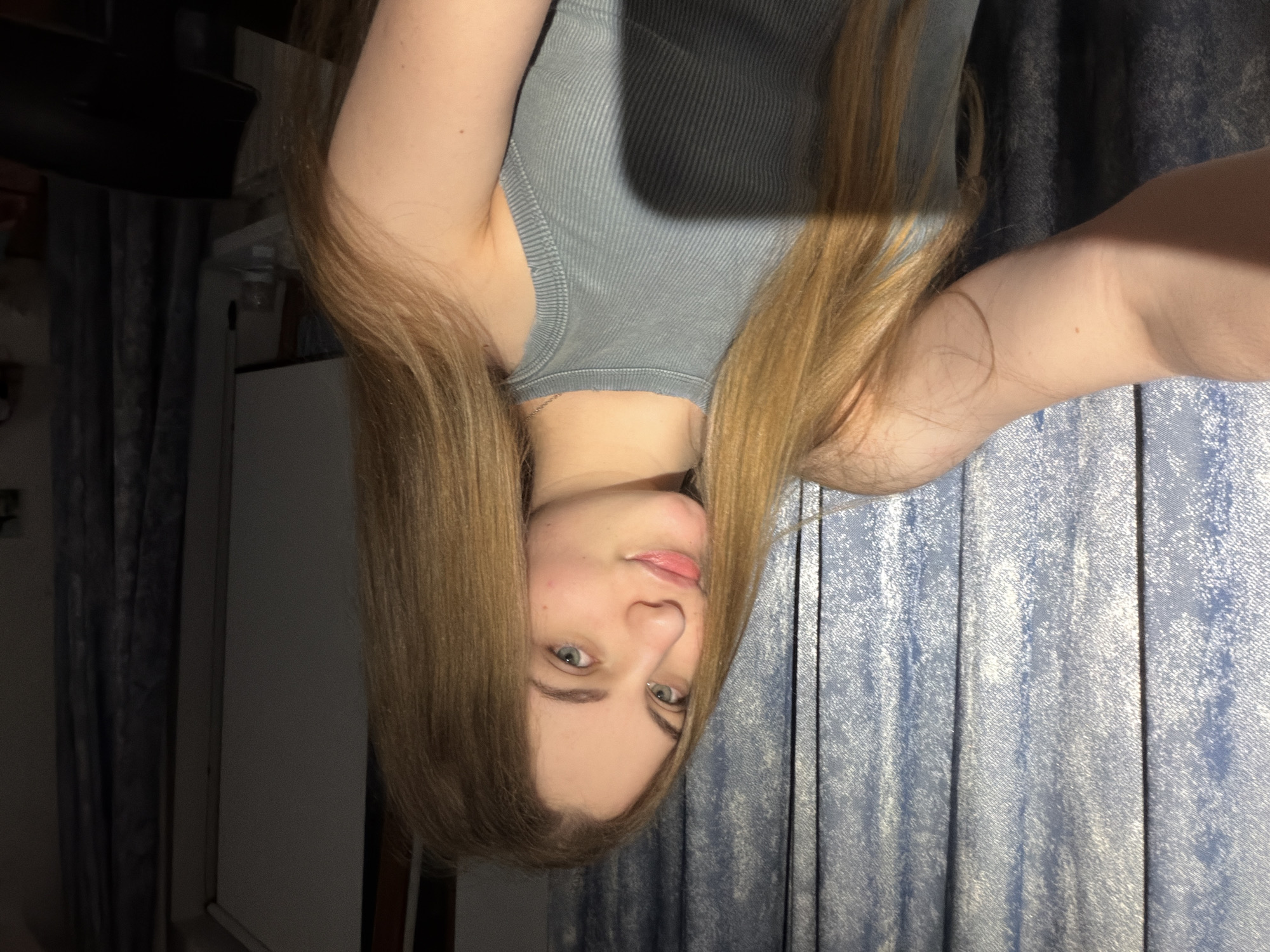 The image is a vertical, close-up, selfie-style photograph of a young woman. She has long, light brown hair that frames her face and falls over her shoulders. Her eyes are a light green or blue, and she is looking directly at the camera with a neutral expression, possibly with a slight smile. She is wearing a grey, ribbed tank top.
The background appears to be an indoor setting. To the left, there are dark blue curtains, and to the right, there is a textured blue and white surface, possibly a wall or a piece of furniture. There is also a glimpse of what looks like a white cabinet or dresser on the left side of the image. The lighting suggests it is daytime, with a light source casting shadows. The overall mood is casual and personal.
Given the location context of Dnipro, Ukraine, and the general style of the photograph, it is likely a personal snapshot taken in a home environment. There is no visible text in the image.