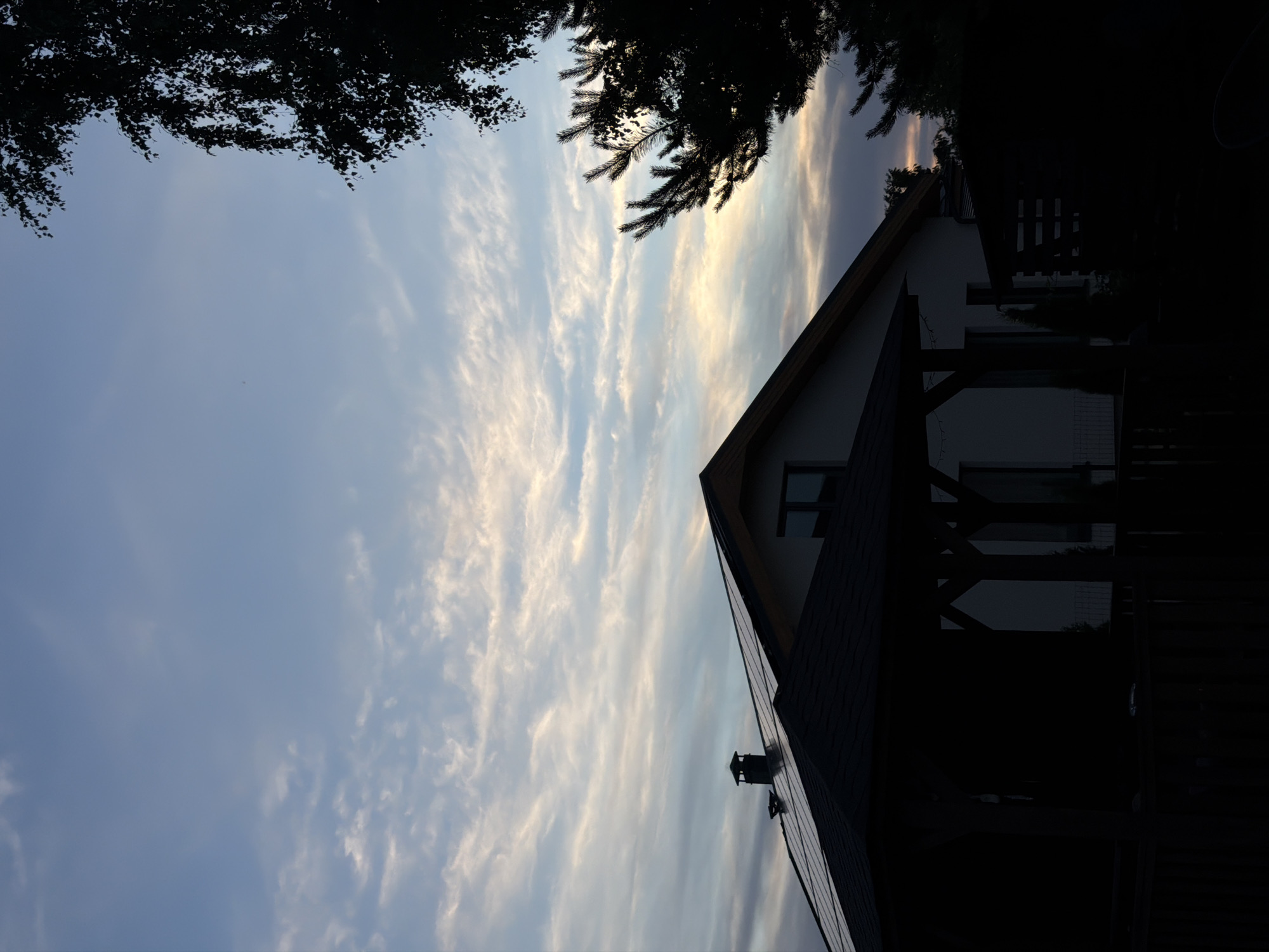 The image shows a silhouette of a house with solar panels on the roof against a cloudy sky. A dark gazebo is in the foreground, obscuring the lower part of the house and garden. Trees are silhouetted on the right side of the frame. The scene is set in the city of Dreiliņi, Latvia.