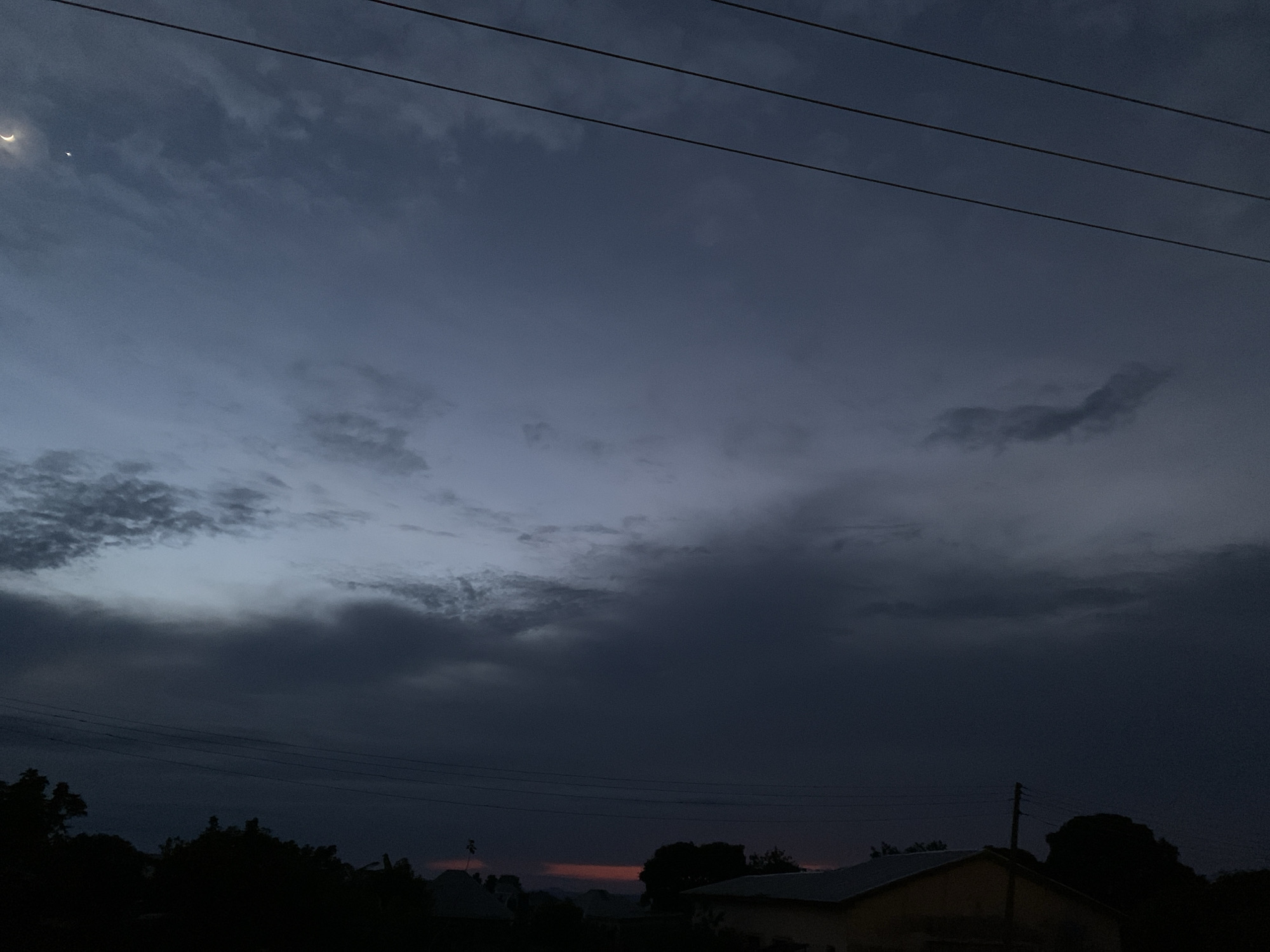 The image shows a dusk sky over Kuru Karama, Nigeria. The sky is a deep blue with scattered clouds, and a sliver of a crescent moon is visible in the upper left corner. Power lines stretch horizontally across the frame. Silhouetted trees and the rooftops of buildings are visible in the lower portion of the image. A faint reddish glow is present on the horizon, suggesting the last vestiges of sunset. The overall impression is one of a peaceful, twilight scene in a semi-urban or rural environment.