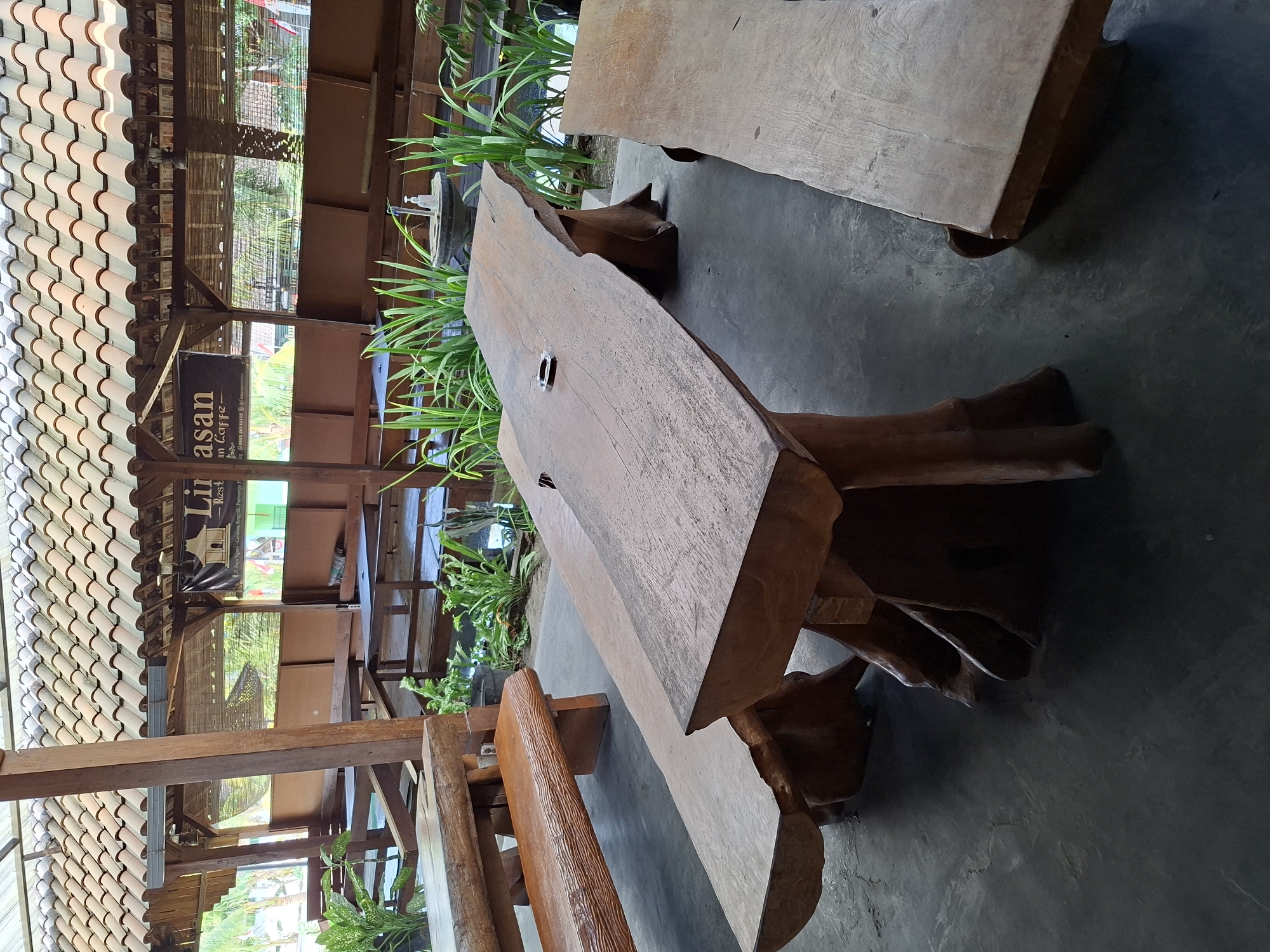The image shows an outdoor seating area at a restaurant in Purworejo, Indonesia. The area features rustic wooden furniture, including a large, live-edge wooden table with a natural tree-stump base, and two long wooden benches. In the background, there are more wooden tables and benches, as well as lush green plants. A tiled roof provides shade over the seating area. A banner with the text "Lirasan Caffe" and an address in Purworejo is visible in the background, suggesting this is the location. The ground is paved with concrete. There are no people visible in the image.