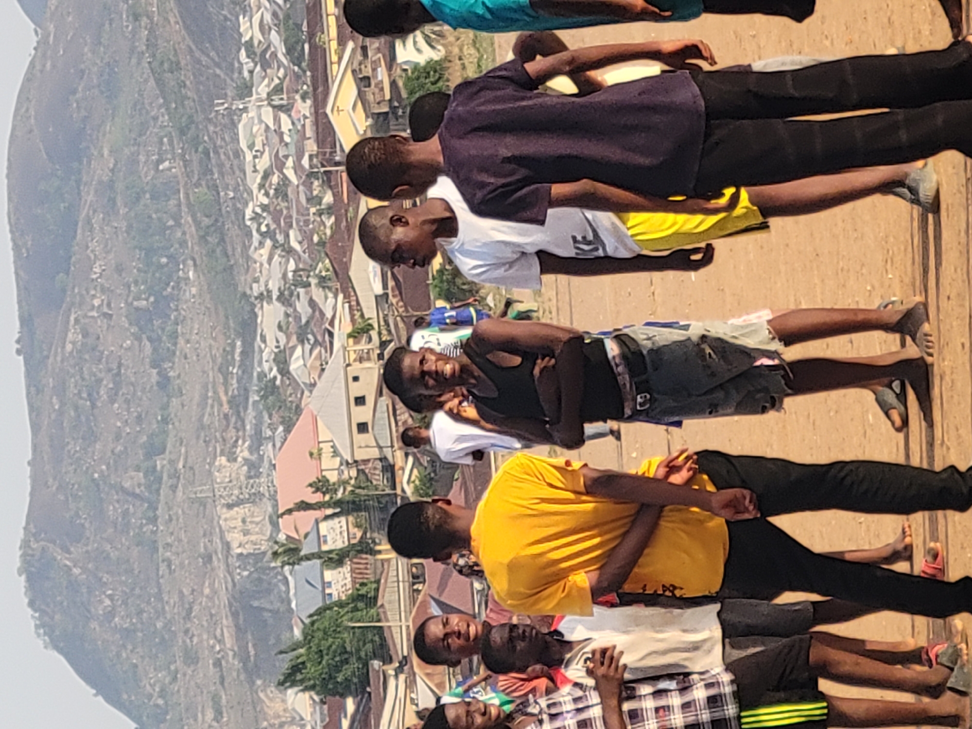 A group of people, primarily young men, are arranged in rows on a dirt surface. Some are lying down while others are seated. The setting appears to be outdoors with a blurred background of buildings and a mountain visible in the distance. The location is Ibo, Nigeria, based on the urban landscape and geographical features in the backdrop. The specific activities or interactions are not readily apparent. The lighting suggests it's daytime.