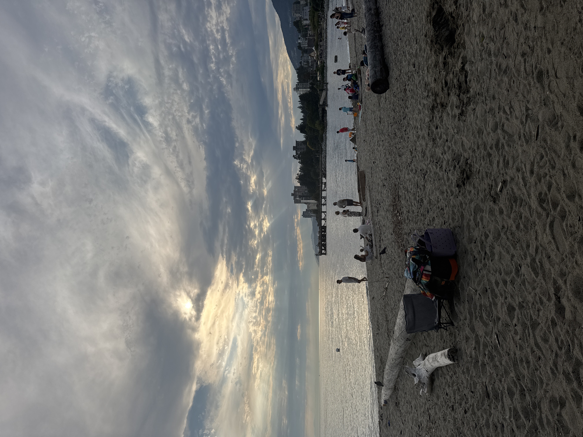 The image depicts a beach scene in West Vancouver, Canada, likely at sunset. The sky is filled with dramatic, cloudy formations with rays of sunlight breaking through, casting a soft light on the water and beach. In the foreground, the sandy beach is covered with scattered driftwood logs, including a large one horizontally positioned across the lower part of the frame. A gray beach chair is set up, accompanied by a pile of colorful beach items, including bags and blankets. Two black birds are perched on the driftwood.
Further back, the calm ocean meets the shore, with a white buoy visible in the water. A pier extends into the water in the middle ground. Along the shoreline, numerous people are scattered, engaged in various activities. Some are standing, some are sitting on blankets, and children appear to be playing. In the background, a line of buildings, some of which are apartment complexes, are nestled amongst trees, with a hillside rising behind them. The overall atmosphere is tranquil and evokes a late afternoon or early evening at the beach.