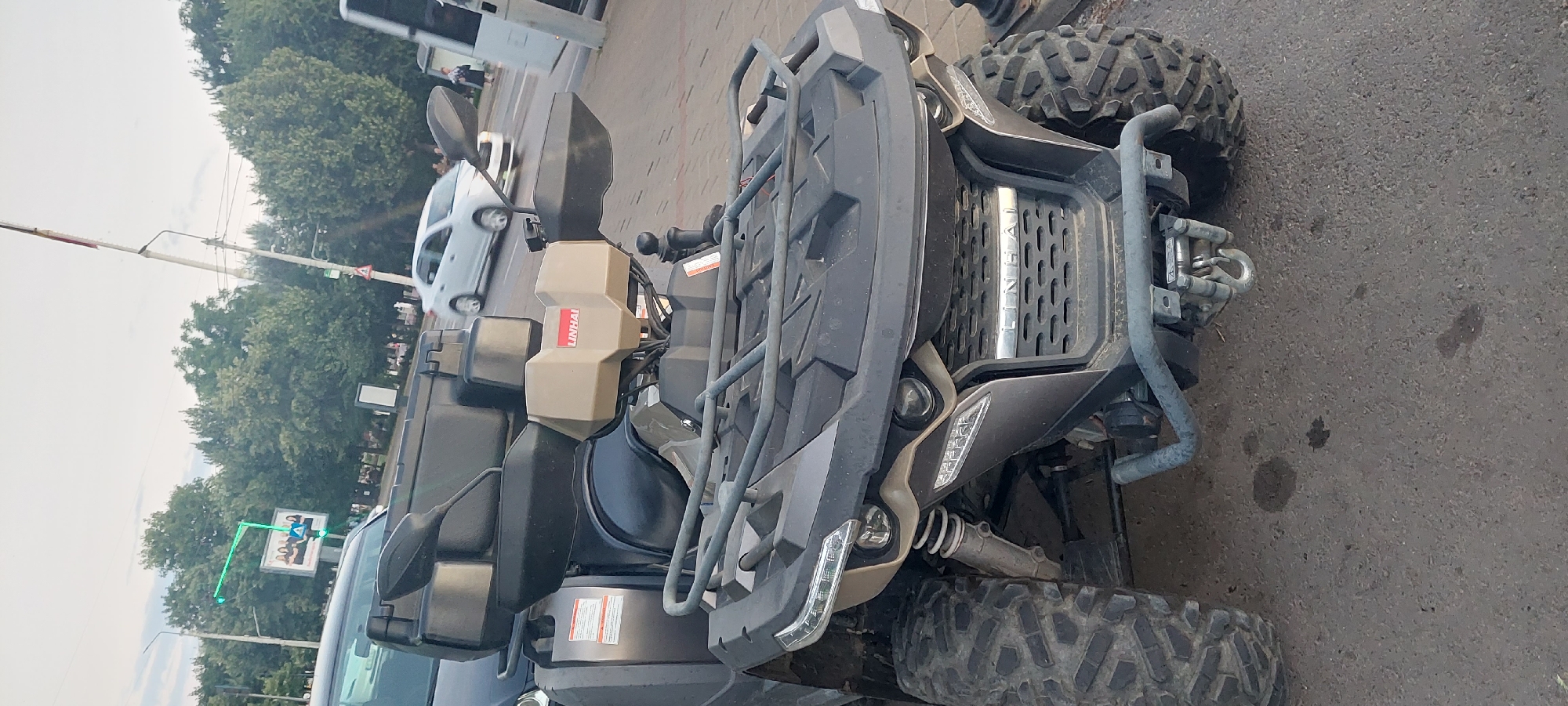 An all-terrain vehicle (ATV) is parked on a paved surface, likely a road or parking lot, in the city of Iași, Romania. The ATV is dark gray with rugged tires. It features a black storage box mounted on the rear, handguards, and a beige housing with "LINHAI" branding on the front. The ATV is equipped with a winch and tow hook. In the background, a white car is driving on the road. Trees line the street, and a traffic light signal is visible. A billboard with a group of people is also present. The sky is overcast.