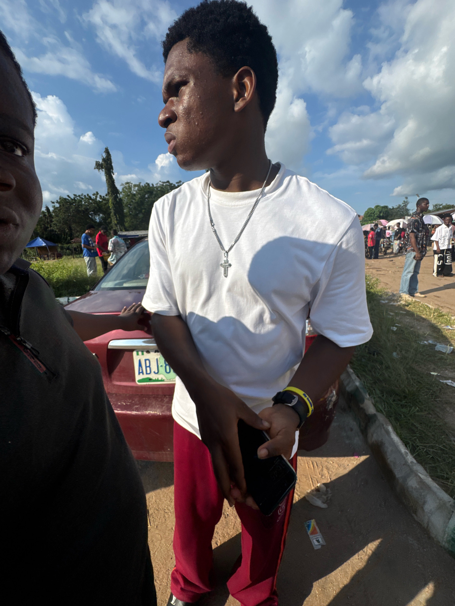 In Ihiagwa, Nigeria, on a bright, cloudy day, two young men are the central figures in the foreground. The man on the right, facing away from the camera and looking to his left, wears a white t-shirt with a silver cross necklace and red pants. A black smartwatch adorns his left wrist, and he holds a smartphone in his right hand. To his left and partially obscuring the frame is another young man, facing the camera and looking towards the viewer. He is dressed in a dark shirt. Behind them, a red car with a Nigerian license plate reading "ABJ-8" is parked. In the background, several other people are visible, some standing near what appears to be a market or gathering, with tents and motorcycles present. The environment is dusty with patches of green grass and trees under a sky filled with scattered clouds.