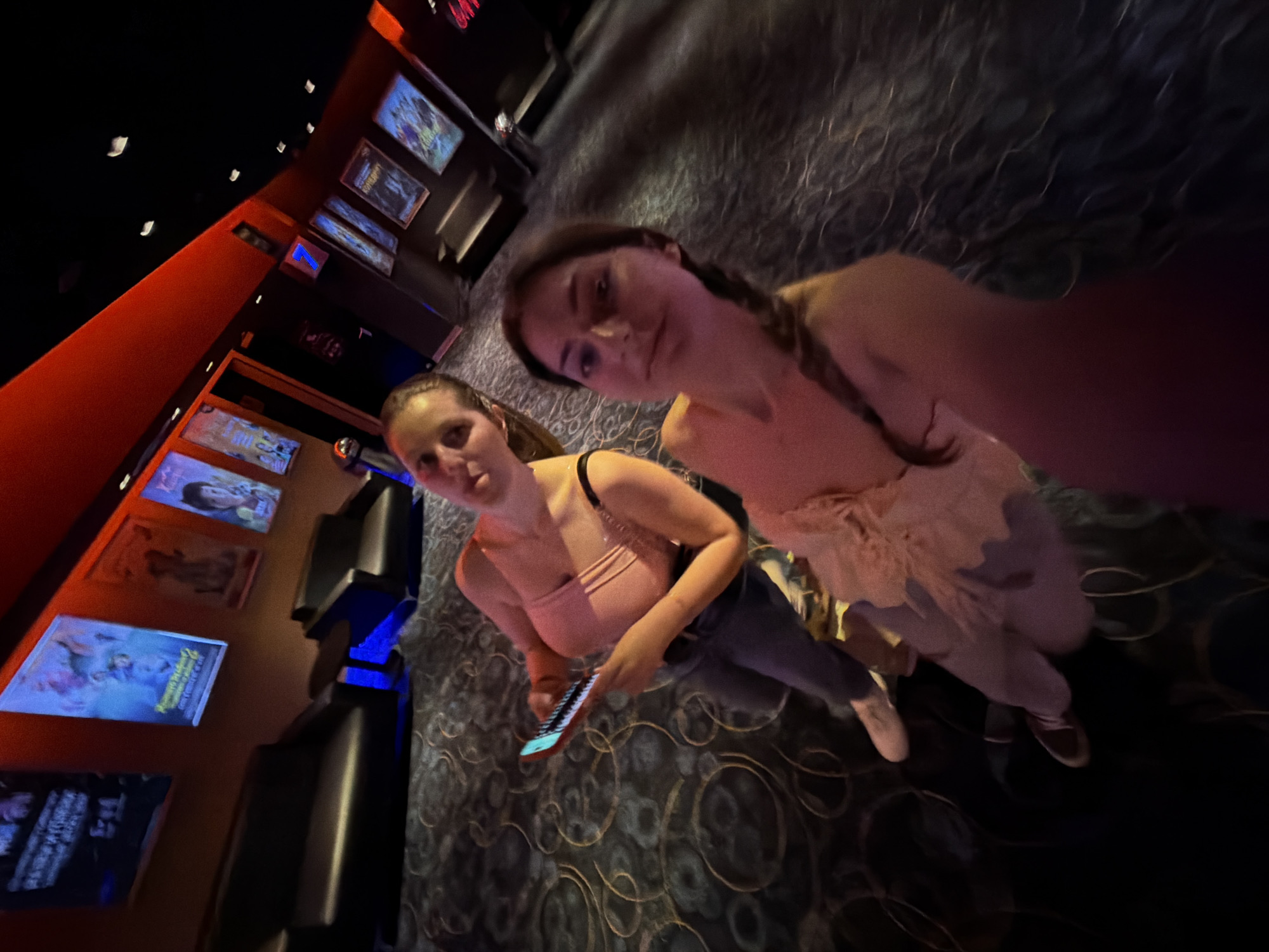 This high-angle, slightly distorted photograph depicts two young women in what appears to be a dimly lit cinema lobby or hallway in Burgas, Bulgaria. The setting is characterized by dark architectural elements, including a dark ceiling with small lights and dark walls, contrasted by a prominent orange-red wall section on the left. The floor is covered with a dark carpet featuring a distinctive swirling pattern of lighter lines.
In the foreground, on the left, one woman with medium-dark hair wears a light-colored, possibly pink, sleeveless top and dark pants. She holds a smartphone in her right hand and looks towards the camera with a neutral expression. Slightly behind and to the right, the second woman has braided hair and is dressed in a light-colored top or dress with a ruffled detail, paired with light pants and dark shoes. She also faces the camera with a neutral gaze.
Along the orange-red wall, several illuminated movie posters or digital screens are visible, showcasing various film titles and promotional artwork. While specific titles are mostly indistinct due to blur and angle, one poster on the far left clearly features animated characters. Further down the wall, dark cinema seats or benches are arranged, and a blue illuminated arrow sign points to the right. The overall dim, artificial lighting suggests an evening or night-time setting, typical of a cinema environment. The women appear to be posing for a casual photograph.