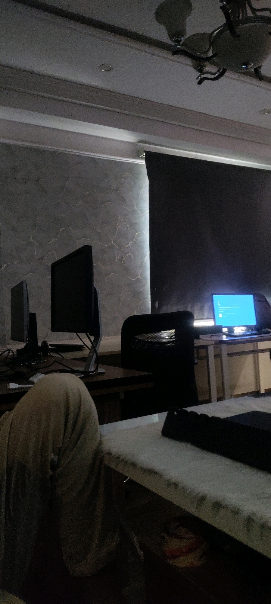 A dimly lit indoor office space, likely in Khana Dak, Pakistan, features two computer monitors on a wooden desk in the foreground. A person, partially visible from the shoulders up, is wearing a beige headscarf or garment, and is seated in front of the monitors. Behind the desk, a black office chair is positioned in front of another desk where a laptop is open and displaying a blue screen with white text. A dark roller blind covers a window or wall behind the laptop. The room has textured, patterned walls and a ceiling with a light fixture. The overall impression is one of a workspace at night or with limited lighting.