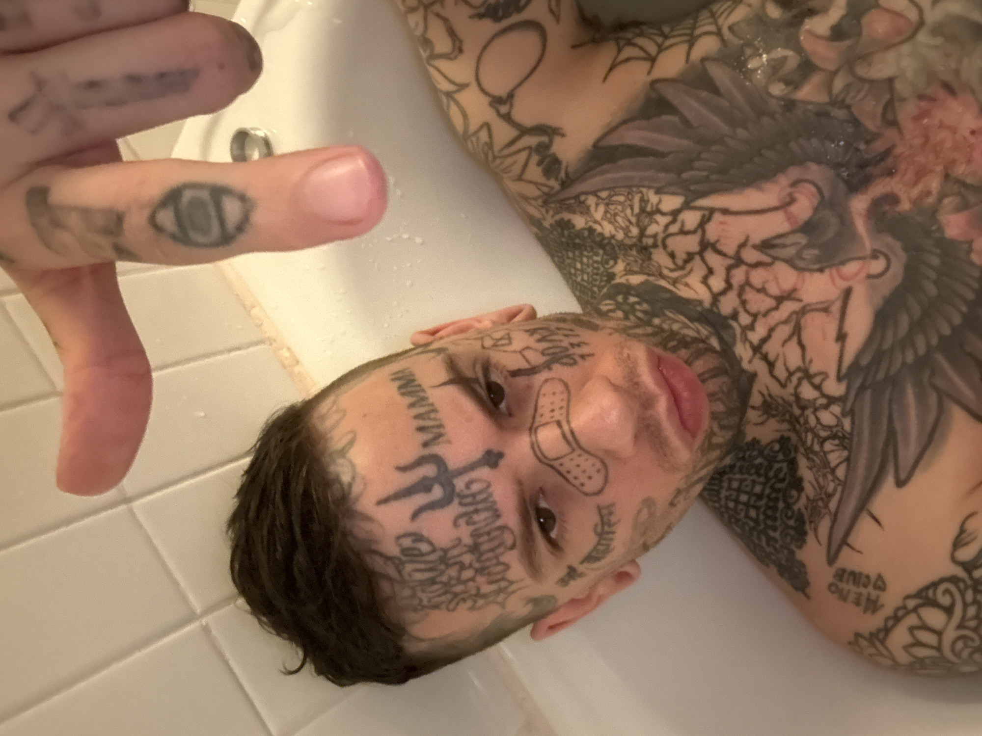 A male individual is depicted in a white bathtub, partially submerged in water, against a backdrop of white tiled walls. The subject has short, dark, wet hair, and facial stubble. His face, neck, chest, shoulders, and fingers are extensively covered with tattoos. Notable facial tattoos include a trident symbol on the forehead, the word "MAMMA," Devanagari script ("सर्वे भवन्तु सुखिनः" - May all be happy) above the right eyebrow, and the numbers "7312" on the right cheek. A tattoo resembling a cartoon band-aid is present on the bridge of his nose. His brown eyes are directed towards the viewer. His right hand is raised into the upper portion of the frame, showcasing finger tattoos, including a circular design on the middle finger and a cross-like symbol on the ring finger. His chest and shoulders feature intricate tattoos, including a large bird resembling an eagle with spread wings, geometric patterns, spiderwebs, and the Italian phrase "MENO DI QUE" (less than that) on his left shoulder. The scene is located in a bathroom in the city of Buenos Aires, Argentina.