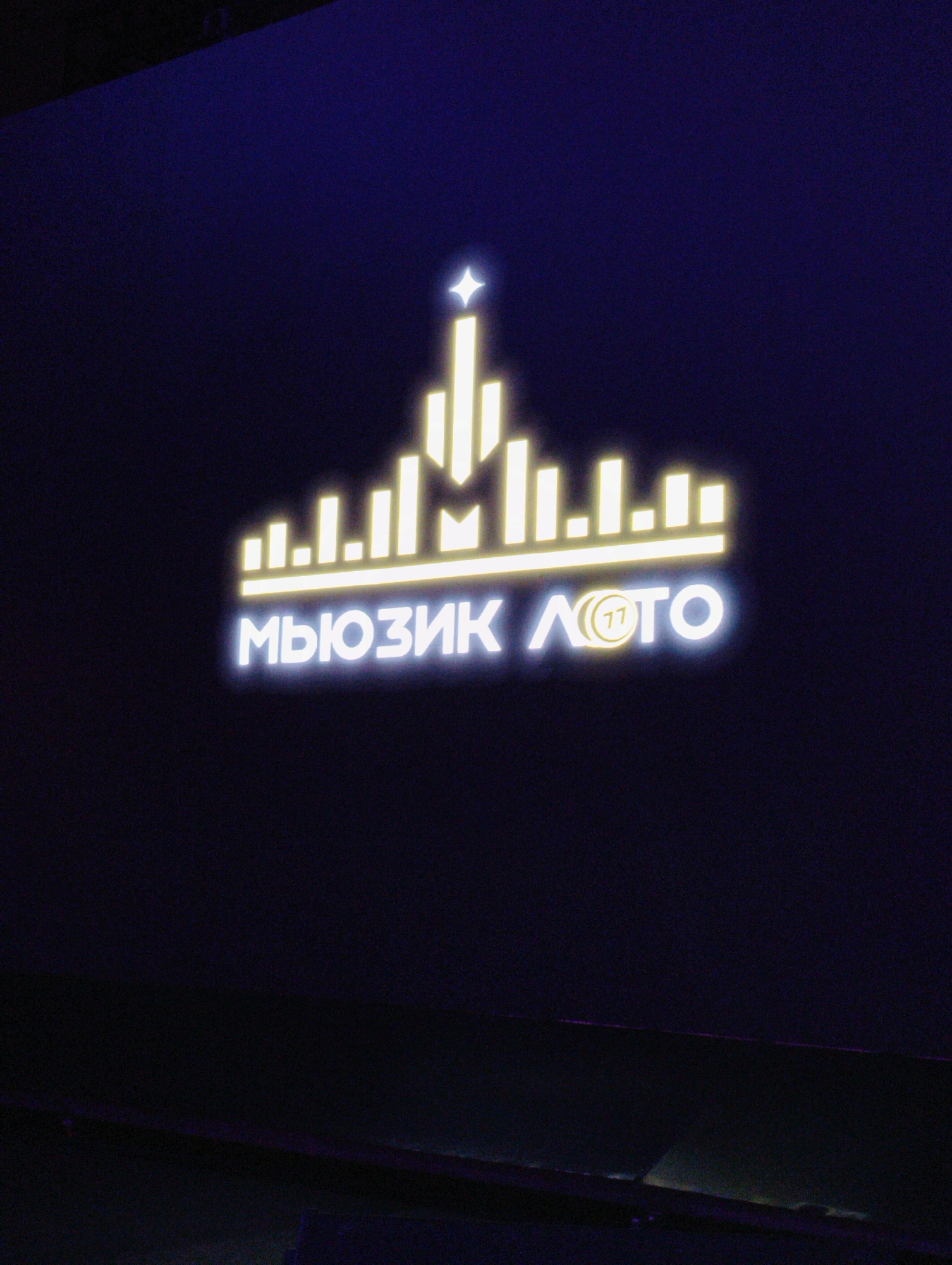 The image features a backlit sign with the Russian words "МЮЗИК ЛОТО" ("MUSIC LOTTO") in white neon lights. Above the words, there's a stylized skyline design composed of vertical bars of varying heights, also in neon yellow lights, with a star at the top center. The background is dark and out of focus. Based on the logo design, it's possible this is located somewhere in Moscow, Russia.