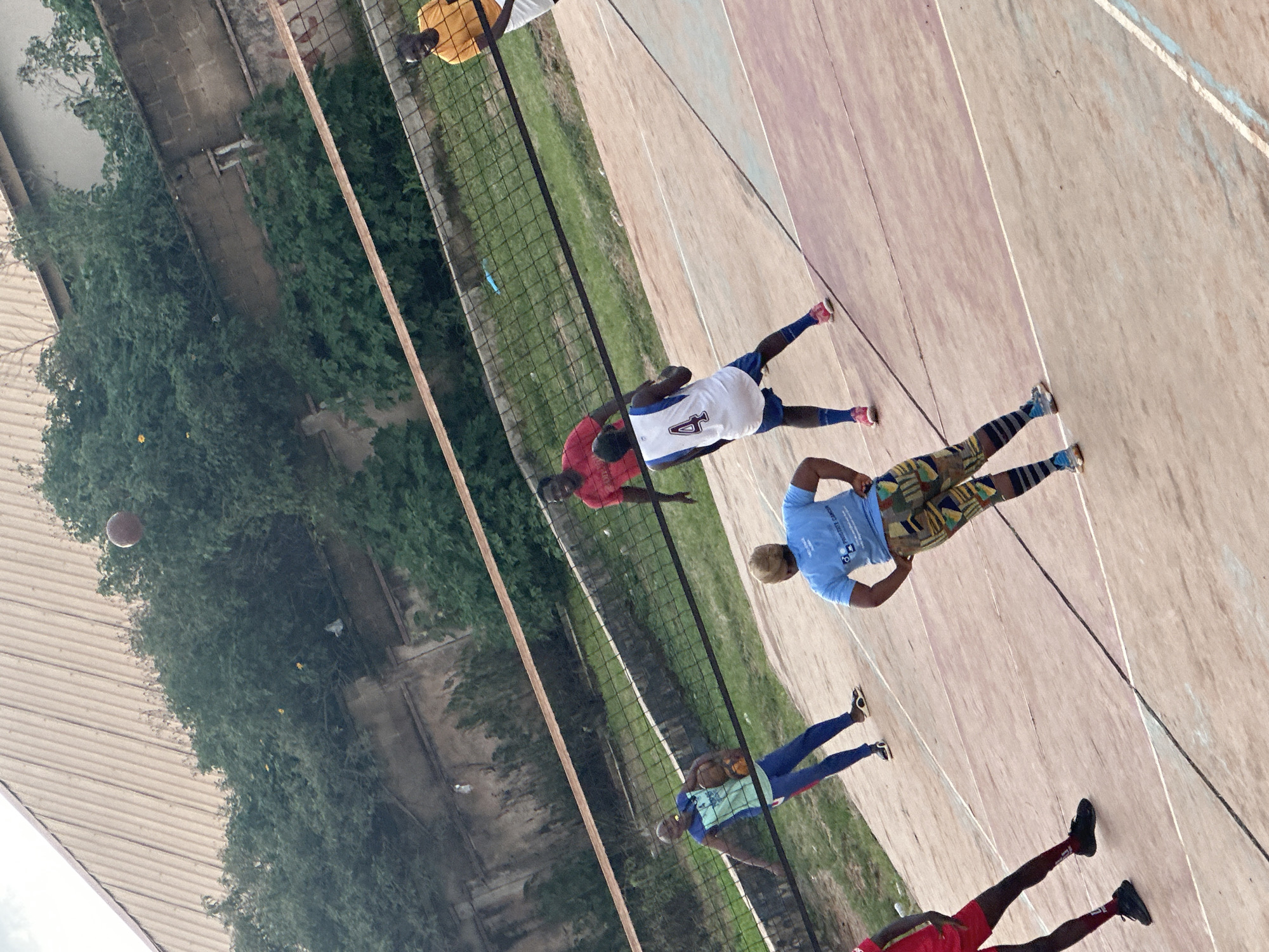 In Gura Top, Nigeria, a group of individuals is engaged in a game of volleyball on an outdoor court. The court surface is a reddish-brown hue with white lines marking its boundaries. A black net divides the court in half, suspended by wooden poles.
Several people are visible on the court. On one side of the net, a person in a blue t-shirt and colorful patterned shorts stands with hands on hips, facing away from the camera. To their left, a person in a blue and teal tank top and blue pants holds a volleyball and appears to be preparing to serve or pass. Another individual in a red t-shirt is also on this side of the net, looking towards the action.
On the other side of the net, a person in a white jersey with the number "4" on the back and blue shorts is positioned in a ready stance, facing the net. To their right, a person in a yellow t-shirt and white shorts smiles, also facing the net. Another person in a red shirt is visible further back on this side.
A brown volleyball is captured in mid-air above the net, suggesting an active play. The background consists of lush green foliage, a concrete wall, and the corrugated metal roof of a building. The lighting suggests it is daytime.