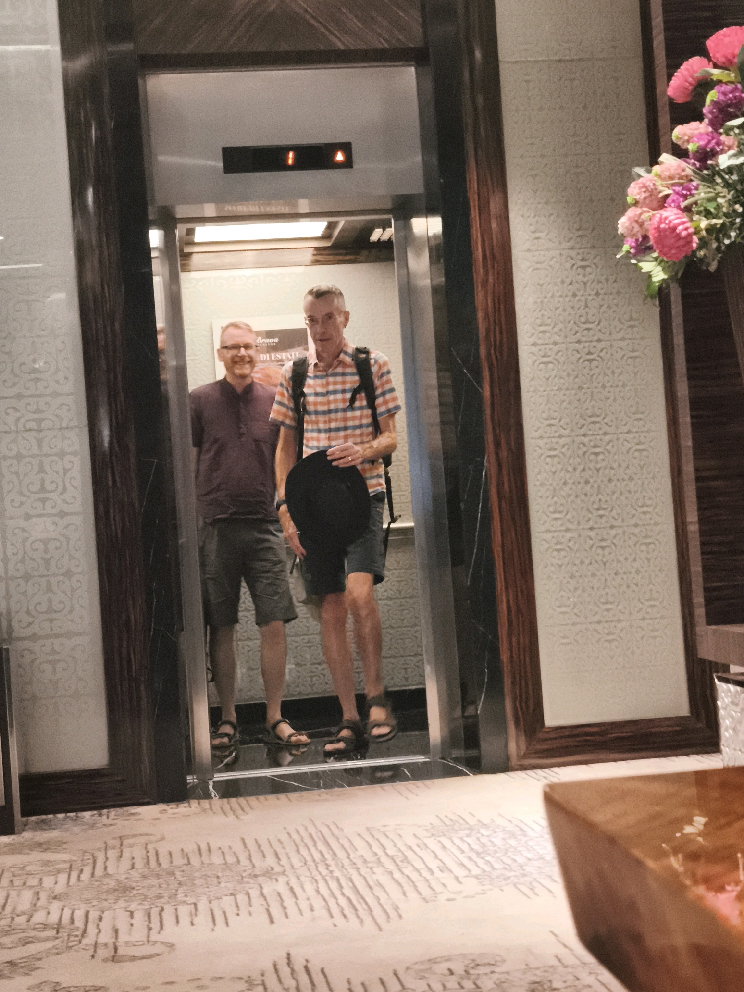 Two men stand in an elevator. The man on the left wears a dark purple polo shirt and khaki shorts. He has glasses and a short beard. The man on the right wears a plaid short-sleeved shirt, dark shorts, and sandals. He also has glasses and is holding a black hat. The elevator has wooden paneling and a reflective floor. The wall behind the men has a textured pattern. The image appears to have been taken in a hotel in Palembang, Indonesia, possibly at the Aryaduta Hotel, given the floral arrangement and interior design visible in the background and foreground.
