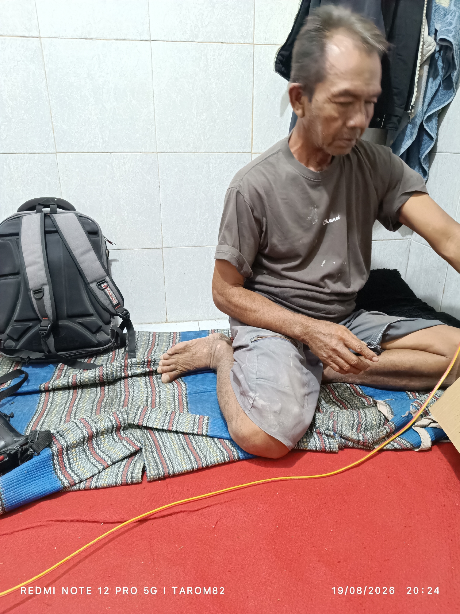 The image depicts an indoor scene with an elderly Asian man sitting on a blue and striped mat on a red floor. He is wearing a gray t-shirt and gray shorts. The man has short, graying hair and appears to be looking down at something in his hands. A black backpack with gray straps is positioned to his left. In the background, there are white tiled walls and some clothing hanging. A yellow cable runs across the foreground of the image. The timestamp "19/08/2026 20:24" and "REDMI NOTE 12 PRO 5G | TAROM82" are overlaid on the bottom of the image. The scene is located in Sidodadi, Indonesia.