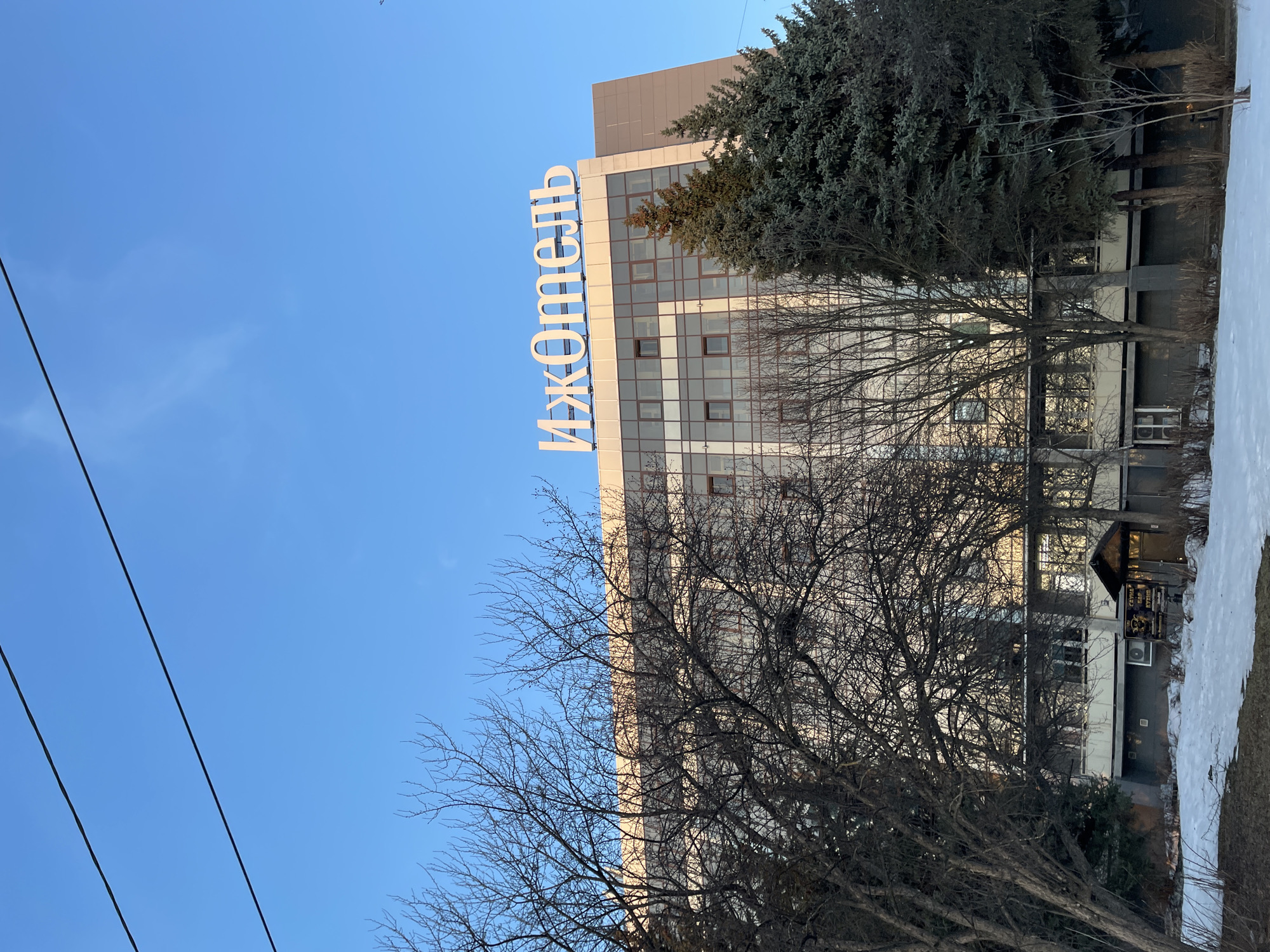The image depicts a multi-story building in Izhevsk, Russia. The building's facade is a light color, possibly beige or white, with a grid-like pattern of windows. A large, vertical sign in Cyrillic script, presumably the name of the building "ИжОтель" (IzHotel), is prominently displayed on the building's edge. Bare trees are in the foreground, partially obscuring the lower floors of the building. A darker green coniferous tree is situated near the top of the building. The sky is a clear, bright blue. Patches of snow are visible on the ground to the right of the building. Power lines cross the left side of the frame.