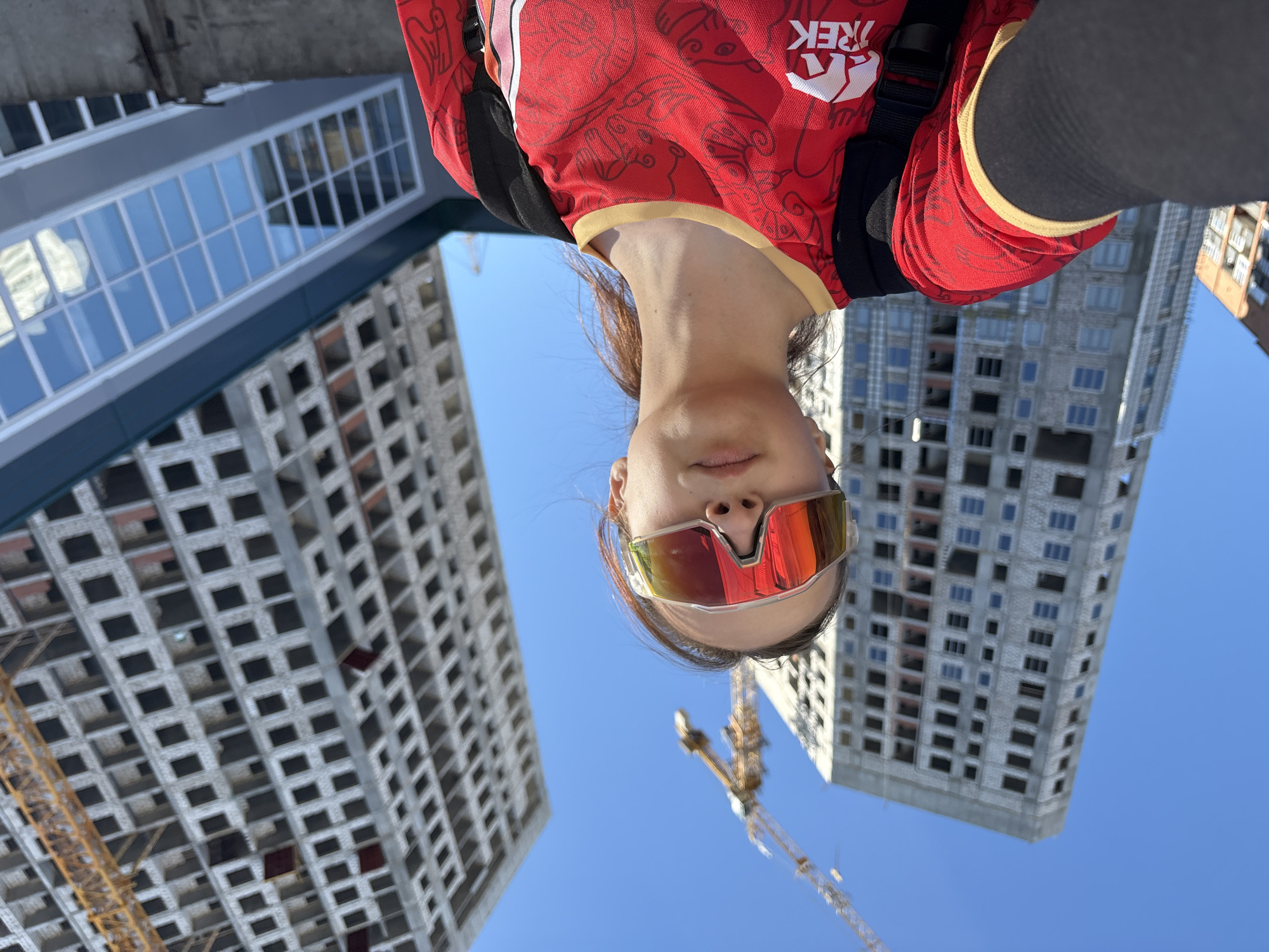 A medium shot captures a woman from a low angle, set against the backdrop of an urban construction site in Perm, Russia. She is oriented upside down in the frame. The woman wears sunglasses with reflective red lenses and a red athletic shirt with a white "TREK" logo on the shoulder. She is likely wearing a backpack. Behind her, two unfinished concrete high-rise buildings under construction loom against a clear blue sky. A yellow construction crane is also visible, indicating ongoing construction activity. The architecture of the buildings is modern, with repetitive window patterns. The scene appears to be taken outdoors during daylight.