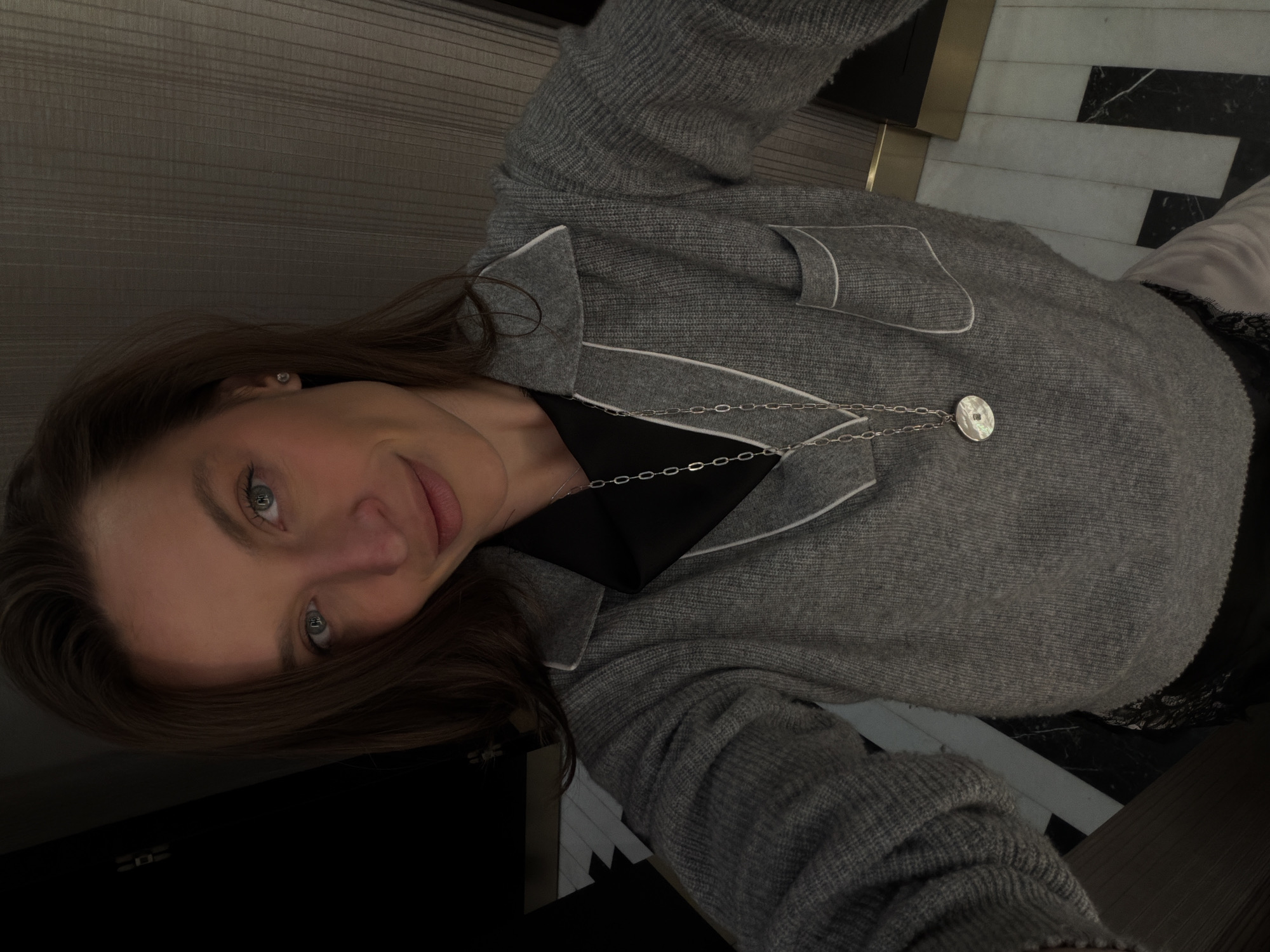 A young woman is facing the camera in an indoor environment. She is wearing a gray cardigan with white trim over a dark top and a silver necklace with a round pendant. Her hair is brown and shoulder-length, and her eyes are light in color. The background shows portions of a room with varied textures and surfaces, including a wall panel on the left and a black-and-white striped floor or wall feature on the right. The precise location within Budva, Montenegro, cannot be determined from the image alone.