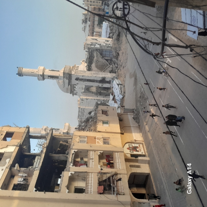 A war-torn cityscape shows extensive building damage and a partially destroyed mosque with a tall minaret. A line of people walks down the street towards the camera. Visible on the side of a building is a faded mural. Debris is scattered throughout the scene. Electrical wires crisscross the sky above the street. The image suggests conflict and devastation.