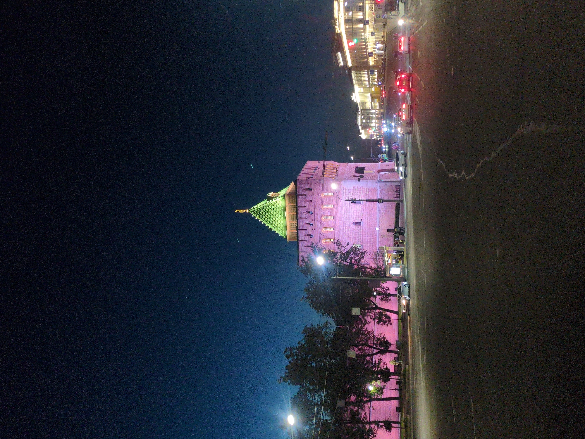 This media file captures a night scene in Nizhny Novgorod, Russia, focusing on a historical building illuminated with pink and green lights. The building appears to be a fortress or tower, with a distinctive green, multi-tiered roof topped with a golden spire. The walls of the structure are bathed in a vibrant pink light, highlighting architectural details such as windows and crenellations.
The scene is set on a street at night, indicated by the dark sky and the artificial lighting. Several cars are visible on the road, their headlights and taillights creating streaks of light, suggesting ongoing traffic. A row of trees lines the sidewalk in front of the building, their leaves also reflecting the pink light. Streetlights illuminate the area, casting a glow on the pavement and tree trunks.
In the background, a street with a line of buildings can be seen, also illuminated with warm, bright lights, suggesting a commercial or public area. The overall atmosphere is one of a lively urban night. The weather appears to be clear, as there are no signs of rain or fog. The image conveys a sense of historical grandeur and modern urban vibrancy coexisting at night. No discernible text is visible in the image.