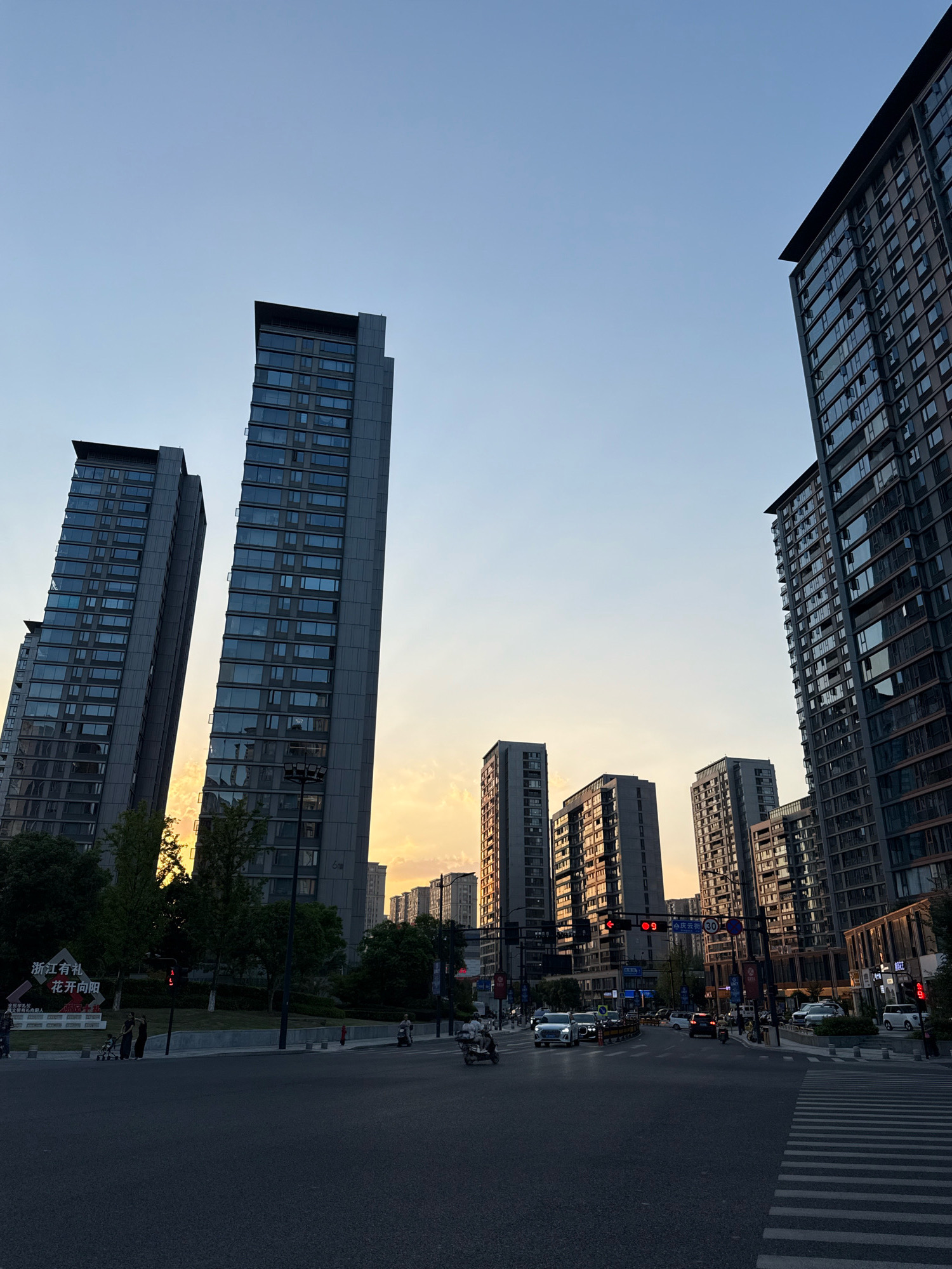 The image depicts a street scene in Yiwu, China, at dusk, characterized by tall residential buildings and active traffic. The sky is a gradient of blue and orange, indicating the setting sun. Several modern, high-rise apartment buildings dominate the skyline, their glass facades reflecting the warm light. In the foreground, a busy intersection with multiple lanes of traffic is visible. Cars and motorcycles are navigating the road, and pedestrian crossings are marked on the asphalt. Streetlights and traffic signals are present. Some pedestrians are seen on the sidewalks, and a few are pushing strollers. Advertisements and signs are displayed on buildings and along the street, some featuring Chinese characters. The overall impression is of a vibrant, urban environment at the end of the day.
